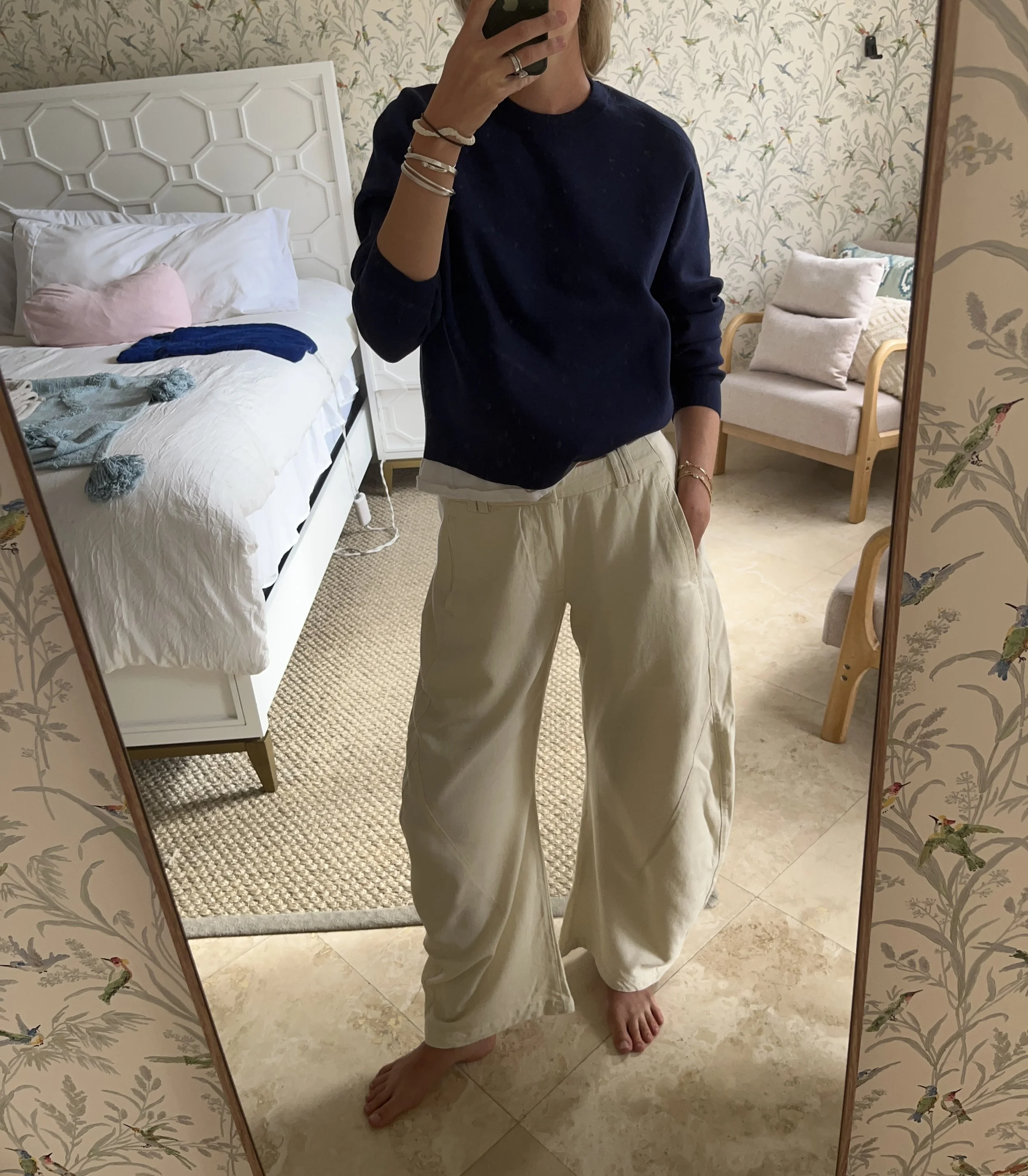 Person taking a mirror selfie in a living room with hardwood floors, wearing a black fleece jacket, gray shirt, black leggings, gray sneakers, and a white cap with a logo.