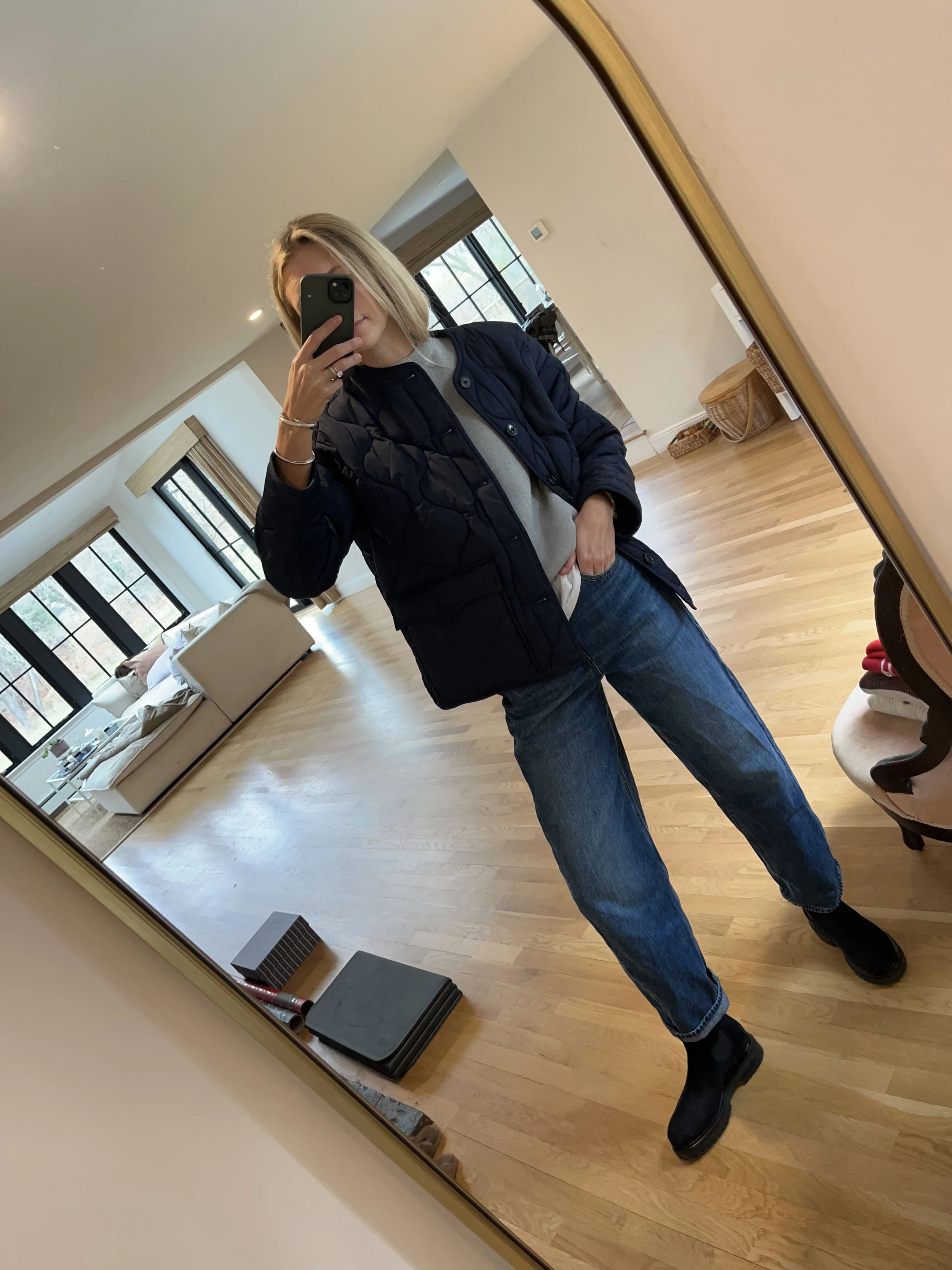 A woman taking a mirror selfie in a spacious living room with wooden floors, large windows, and minimal furniture, wearing a beige jacket, black top, blue jeans, and tan shoes.