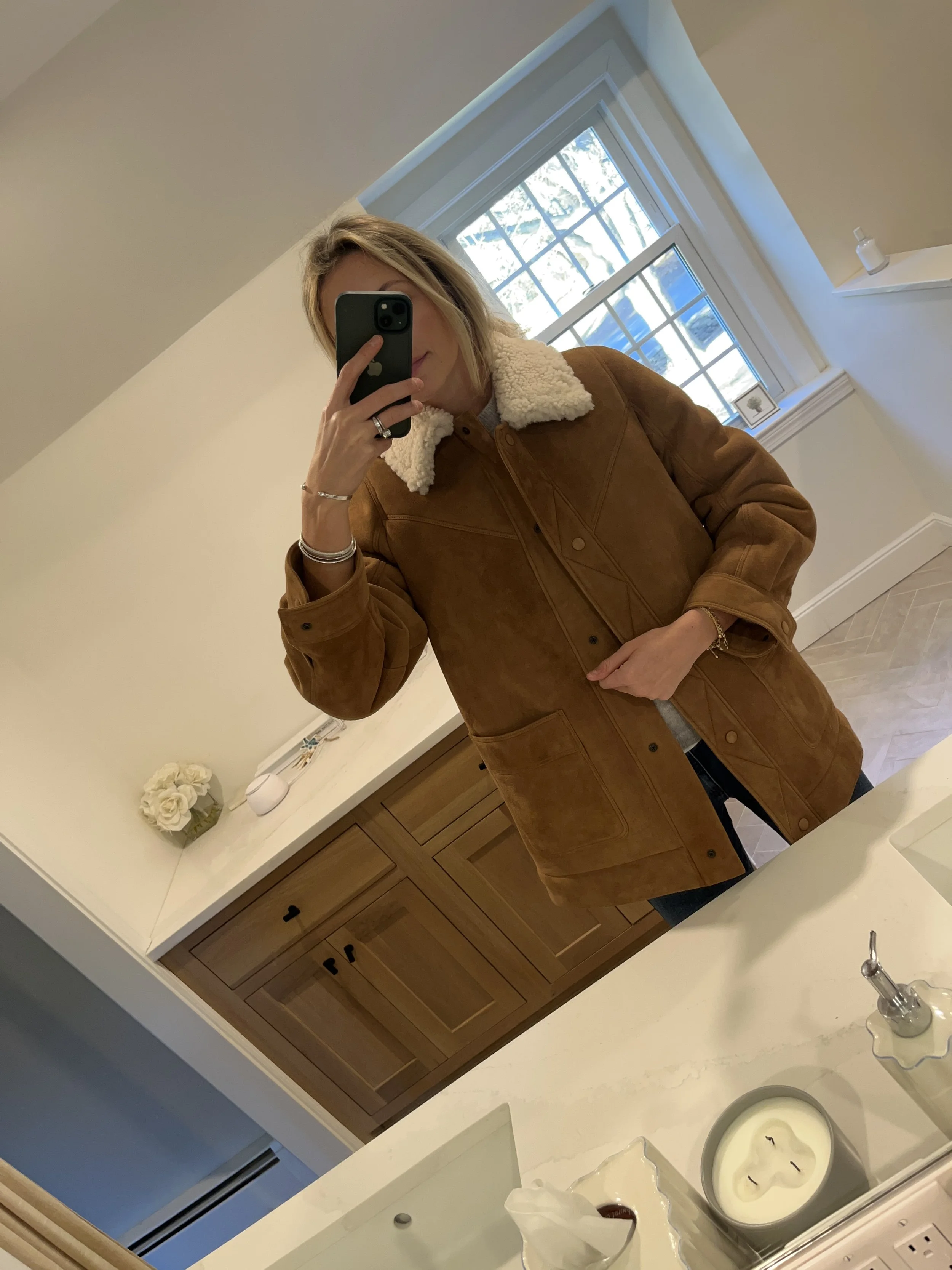 A woman taking a mirror selfie in a spacious, well-lit living room with hardwood floors, large windows, a white sofa, and a side table with a plant.