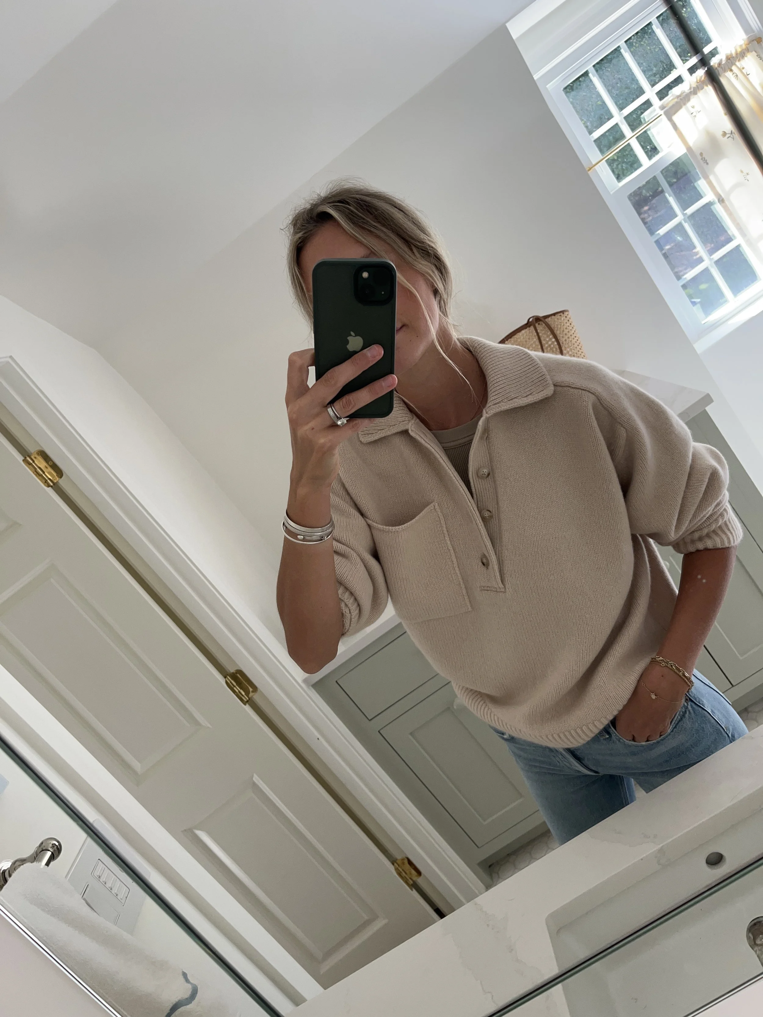 Person taking a mirror selfie in a room with wooden floors, wearing a striped brown and white long-sleeve shirt, light blue jeans, and black and white sneakers.