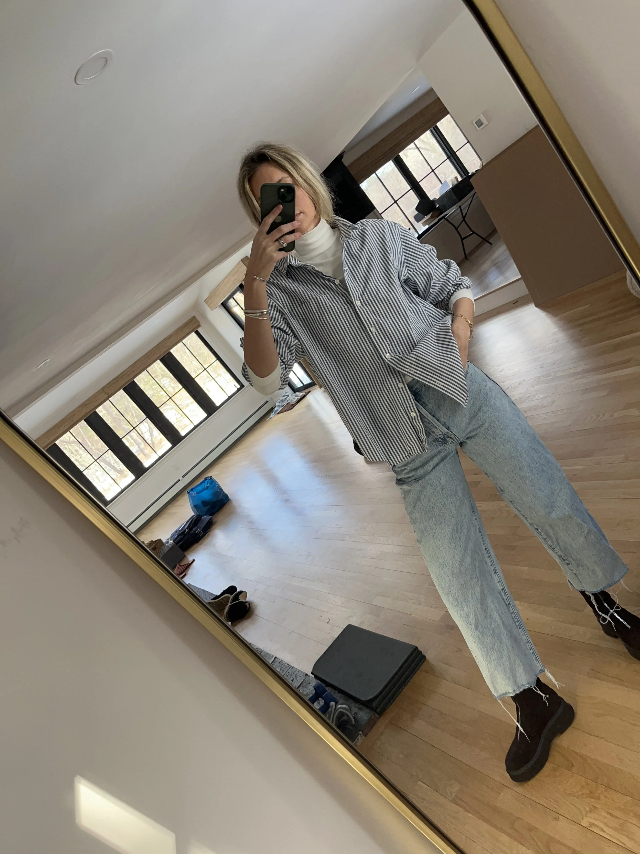Person taking a mirror selfie in a living room with hardwood floors, wearing a black fleece jacket, gray shirt, black leggings, gray sneakers, and a white cap with a logo.