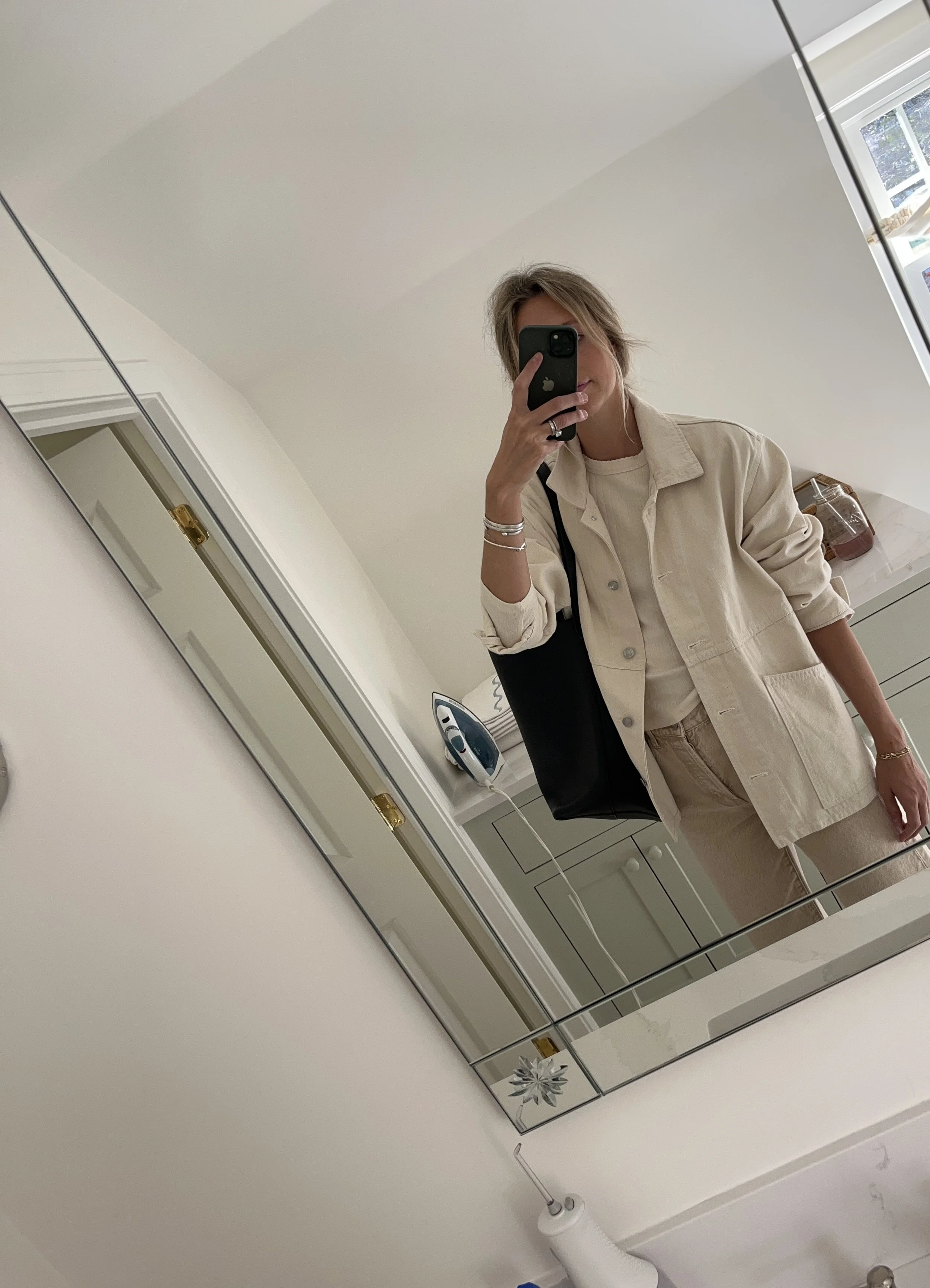 Person taking a mirror selfie in a living room with hardwood floors, wearing a black fleece jacket, gray shirt, black leggings, gray sneakers, and a white cap with a logo.