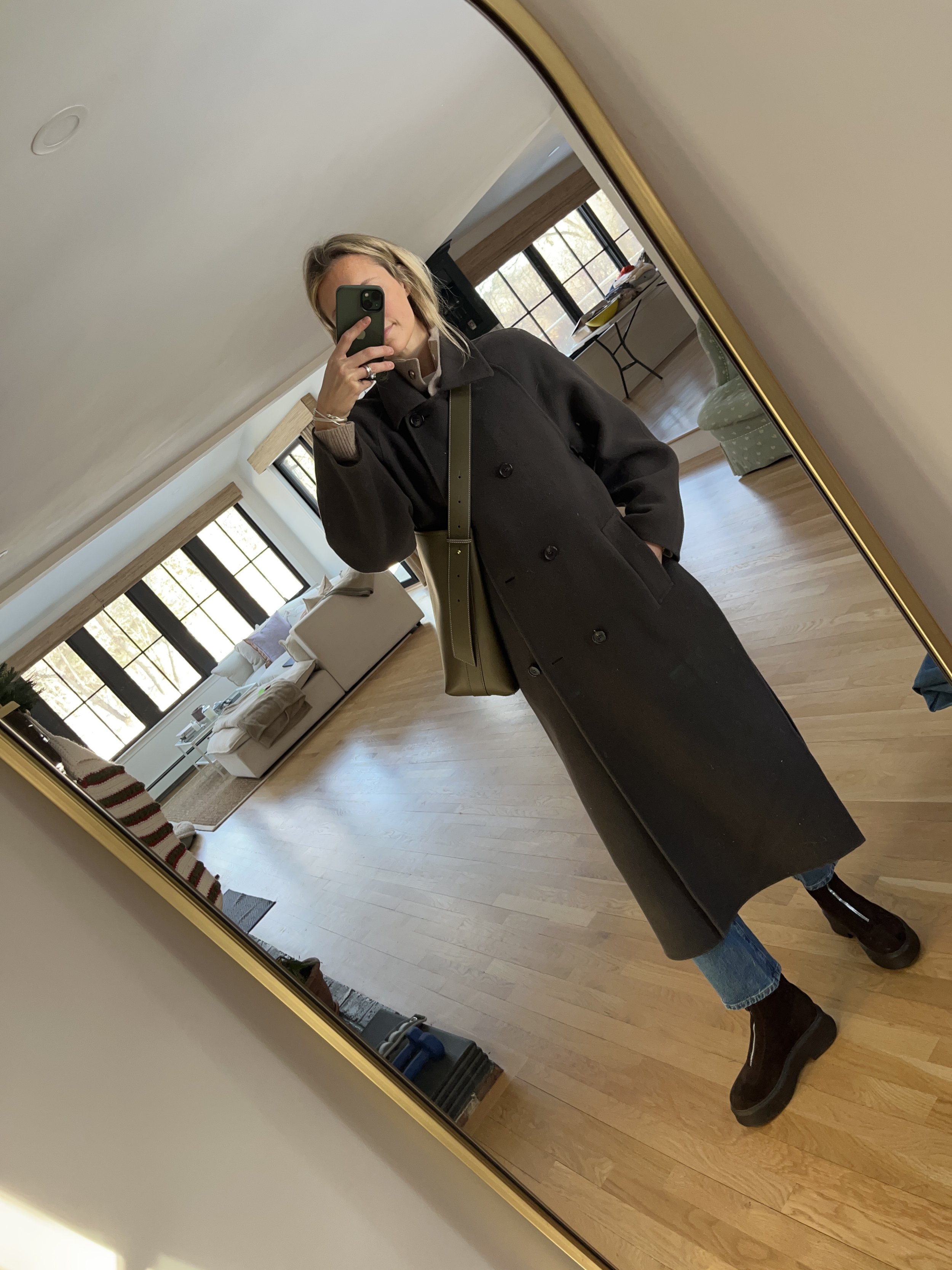 A woman taking a mirror selfie in a spacious, well-lit living room with hardwood floors, large windows, a white sofa, and a side table with a plant.