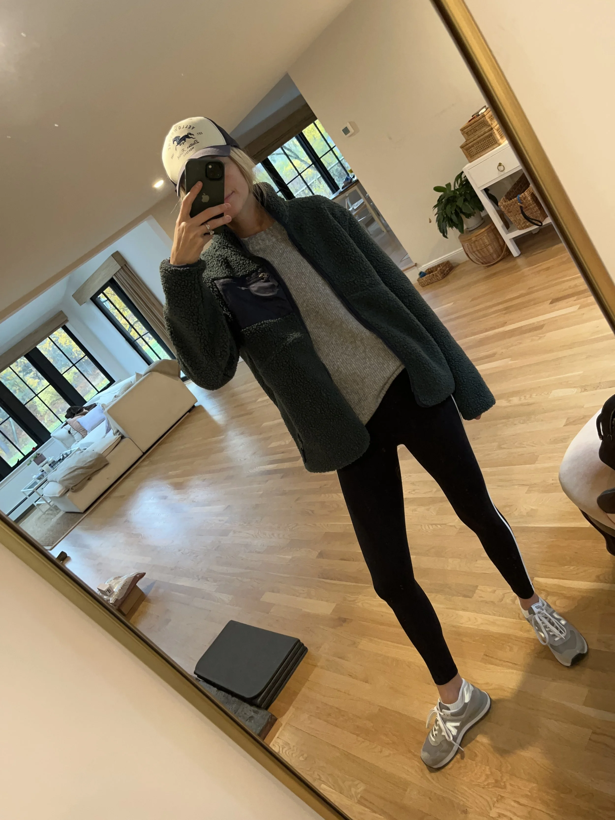 Person taking a mirror selfie in a living room with hardwood floors, wearing a black fleece jacket, gray shirt, black leggings, gray sneakers, and a white cap with a logo.