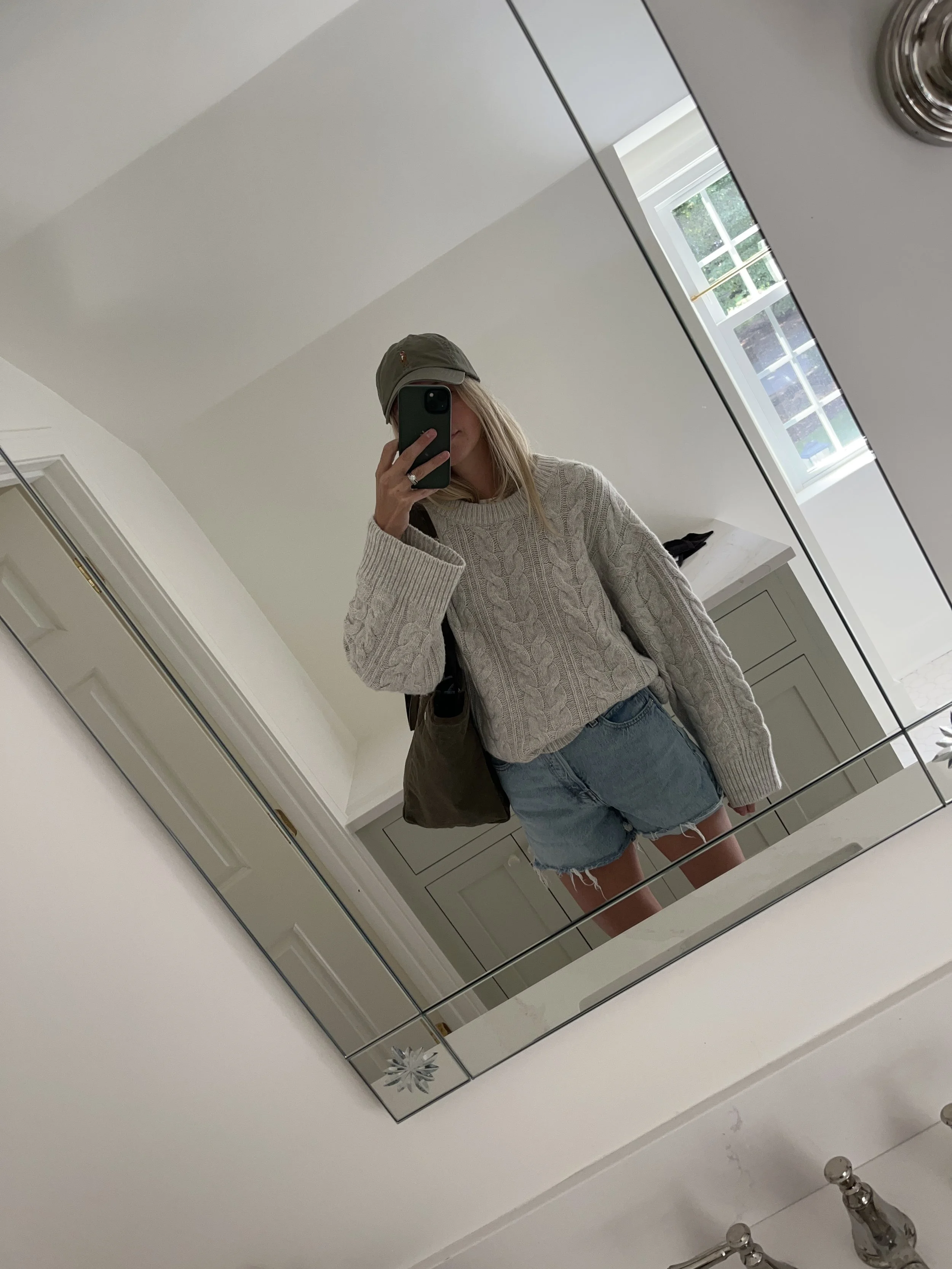 A woman taking a mirror selfie in a spacious living room with wooden floors, large windows, and minimal furniture, wearing a beige jacket, black top, blue jeans, and tan shoes.