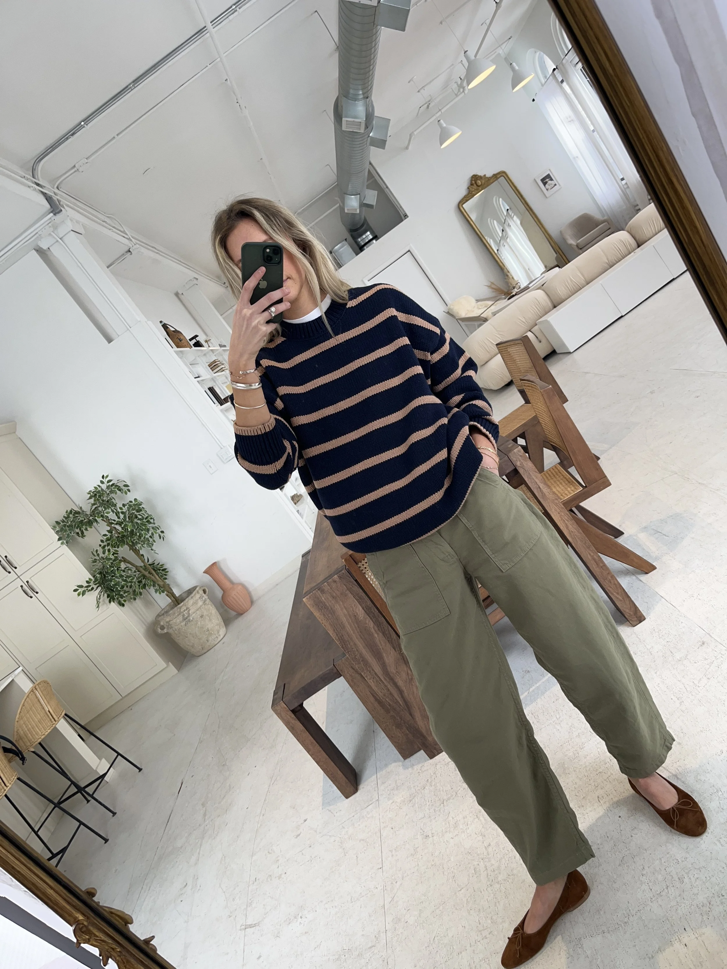 Person taking a mirror selfie in a room with wooden floors, wearing a striped brown and white long-sleeve shirt, light blue jeans, and black and white sneakers.