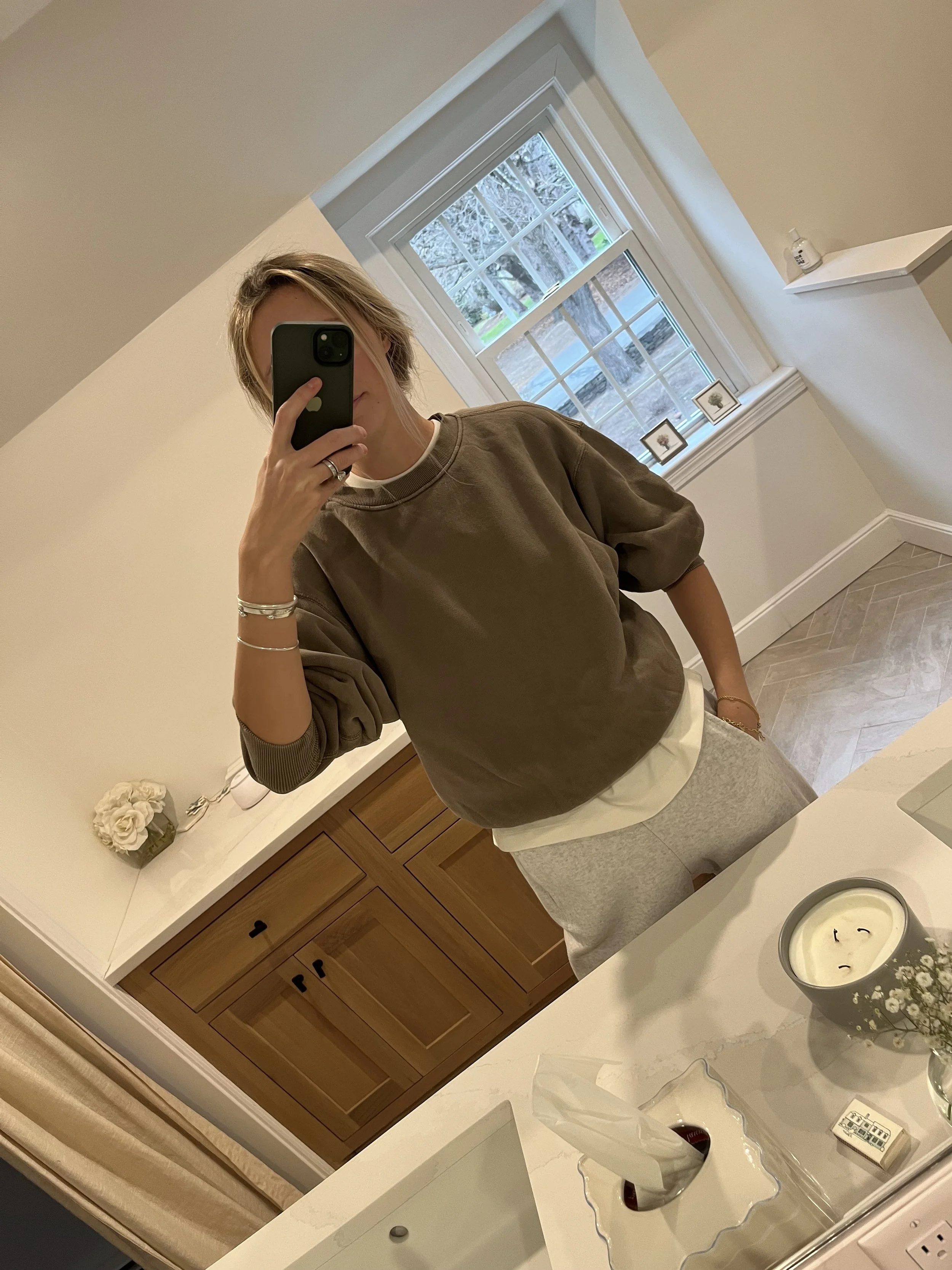 Person taking a mirror selfie in a room with wooden floors, wearing a striped brown and white long-sleeve shirt, light blue jeans, and black and white sneakers.