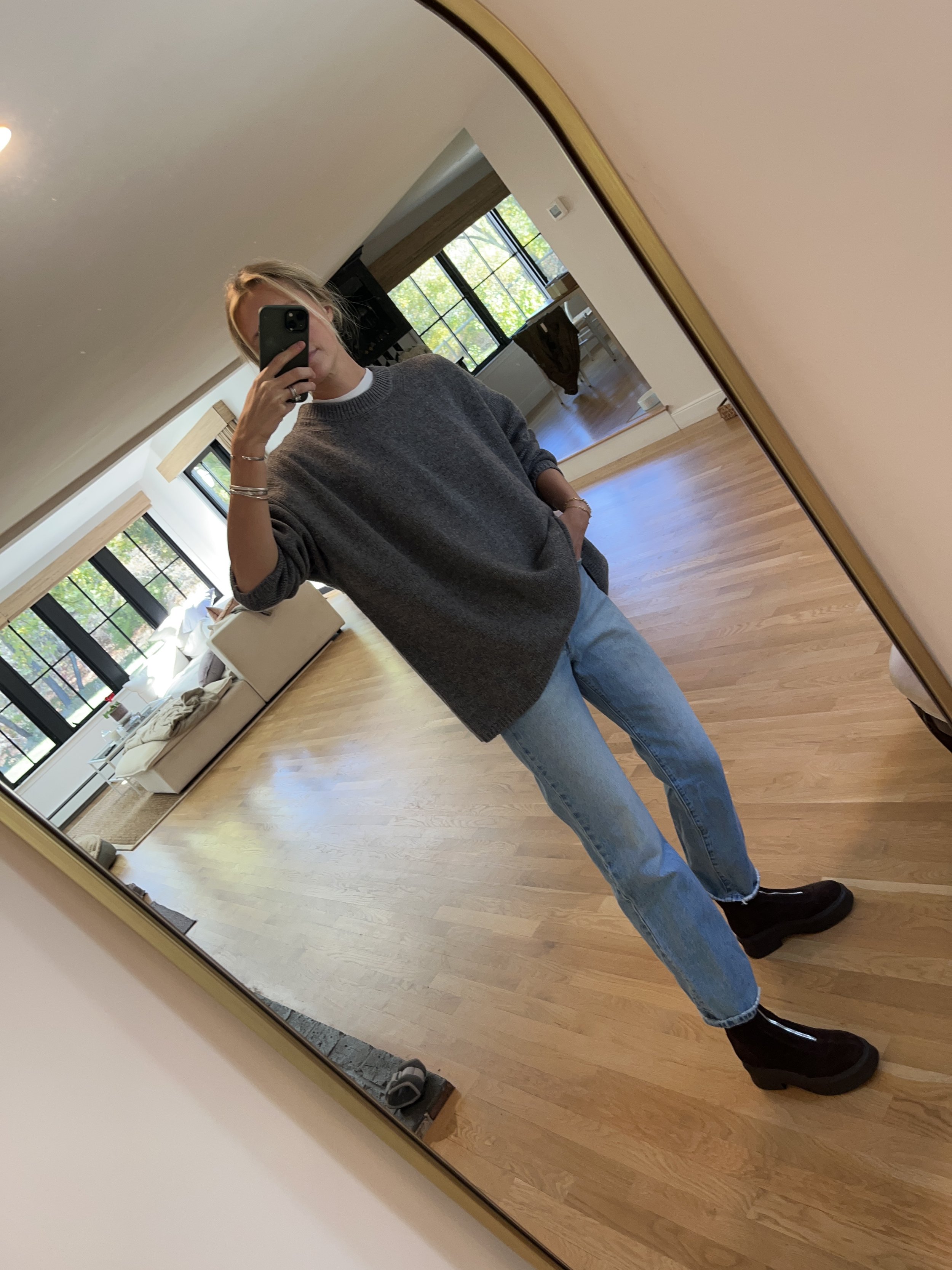 Person taking a mirror selfie in a bright living room, wearing a gray sweater, light blue jeans, and black boots.