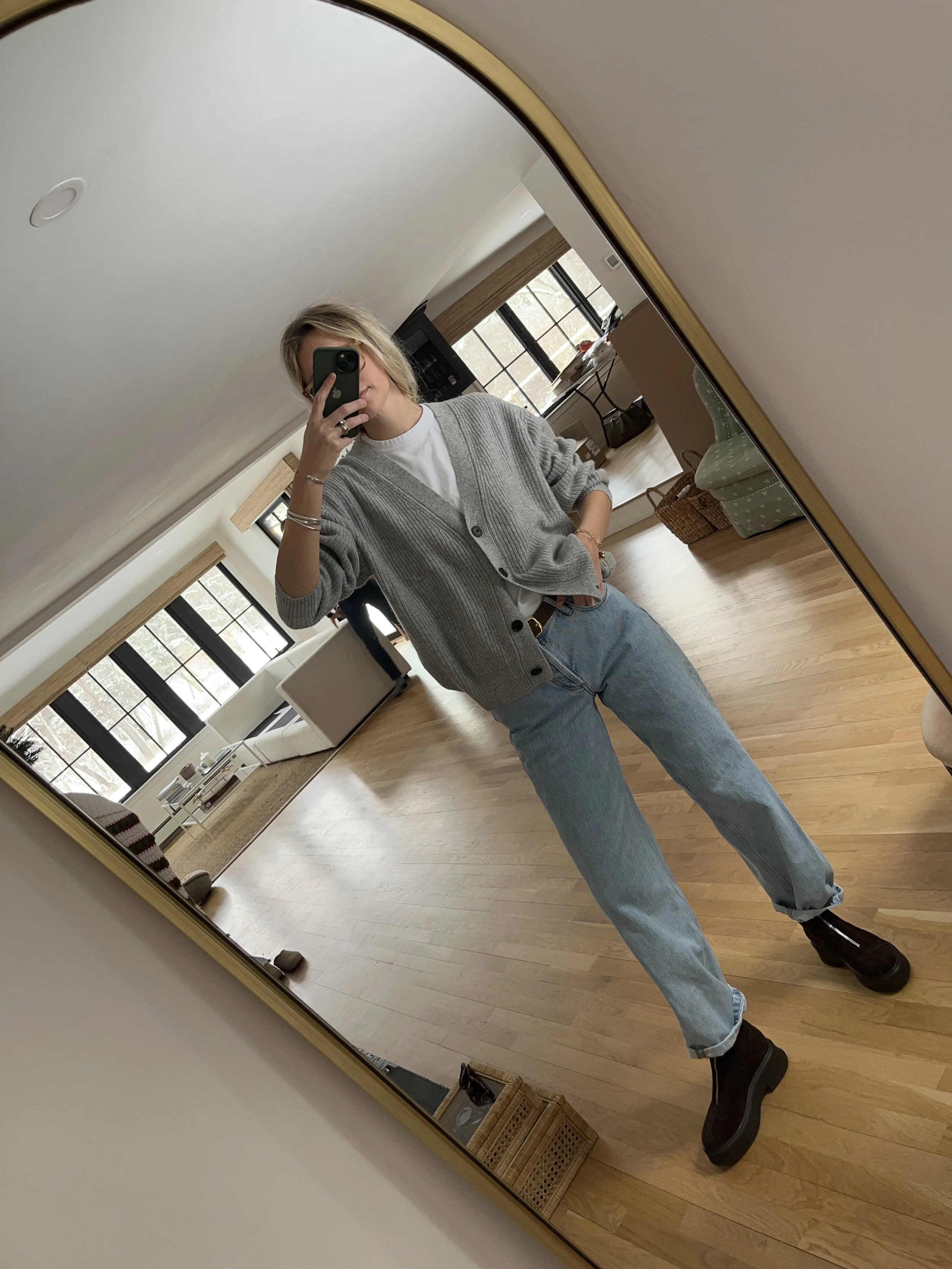 Person taking a mirror selfie in a living room with hardwood floors, wearing a black fleece jacket, gray shirt, black leggings, gray sneakers, and a white cap with a logo.