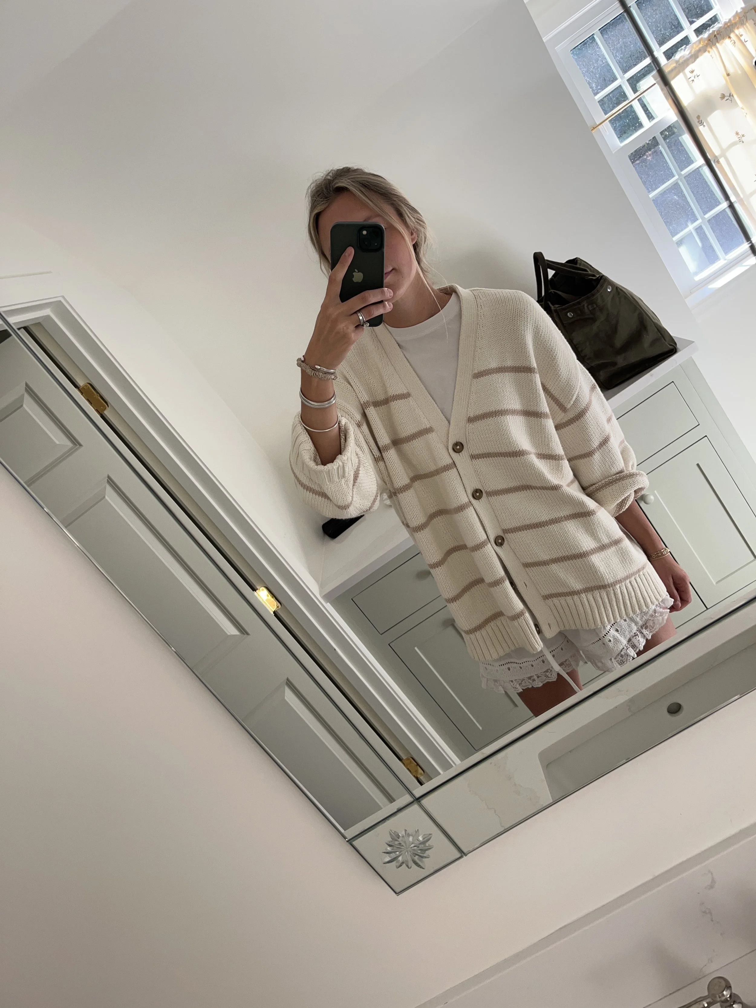 Person taking a mirror selfie in a room with wooden floors, wearing a striped brown and white long-sleeve shirt, light blue jeans, and black and white sneakers.