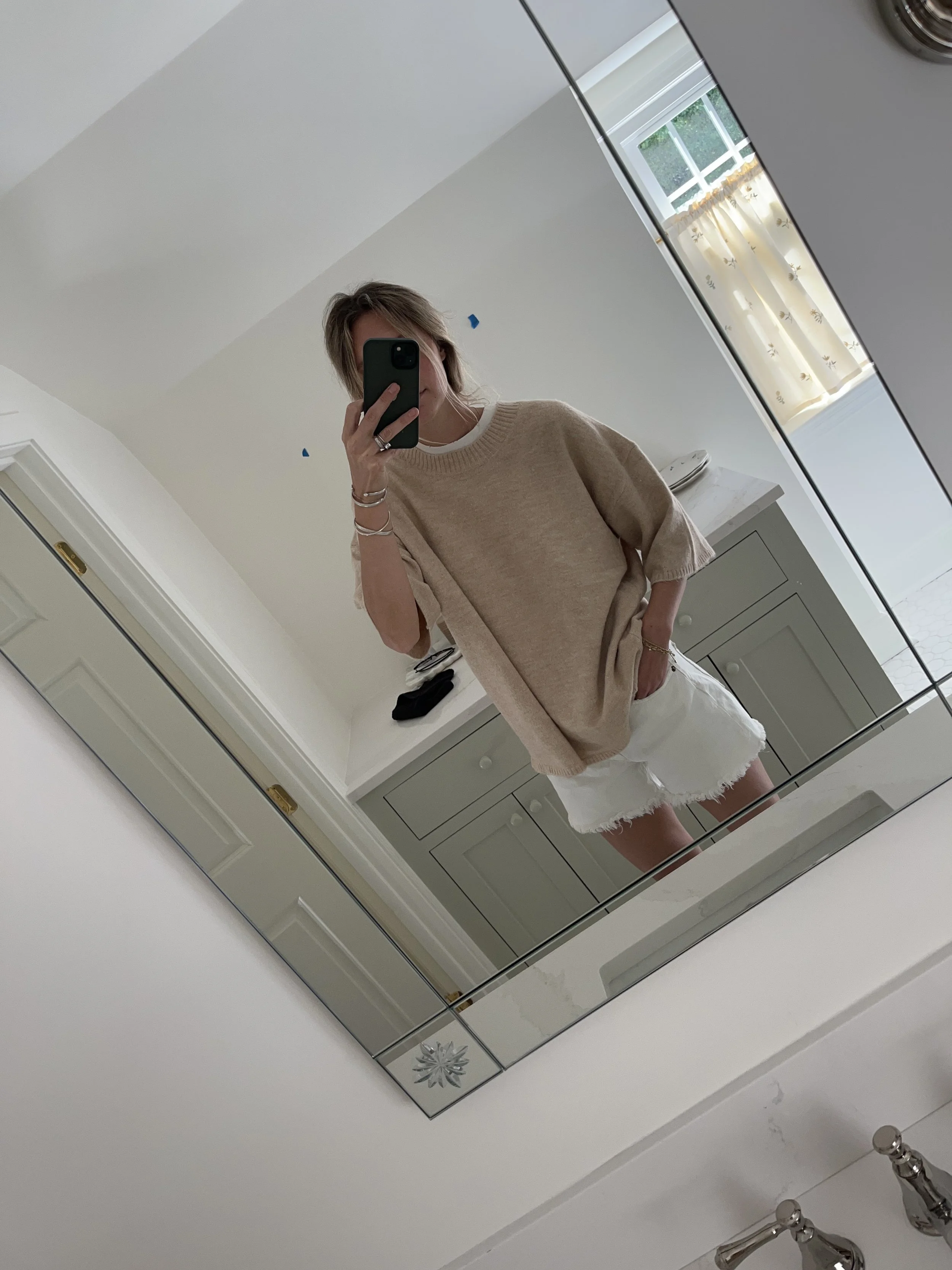 Person taking a mirror selfie in a room with wooden floors, wearing a striped brown and white long-sleeve shirt, light blue jeans, and black and white sneakers.