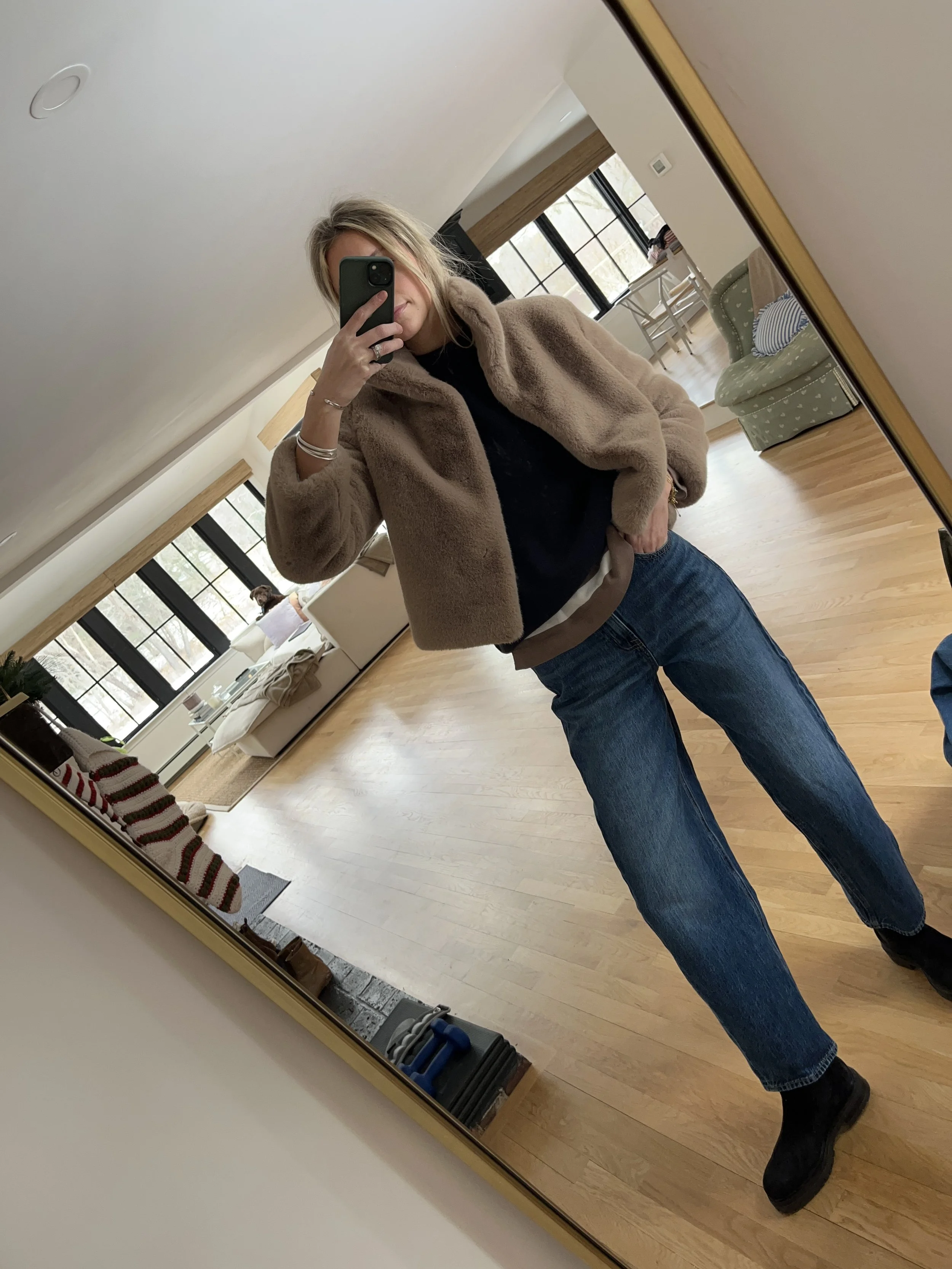 A woman taking a mirror selfie in a spacious living room with wooden floors, large windows, and minimal furniture, wearing a beige jacket, black top, blue jeans, and tan shoes.