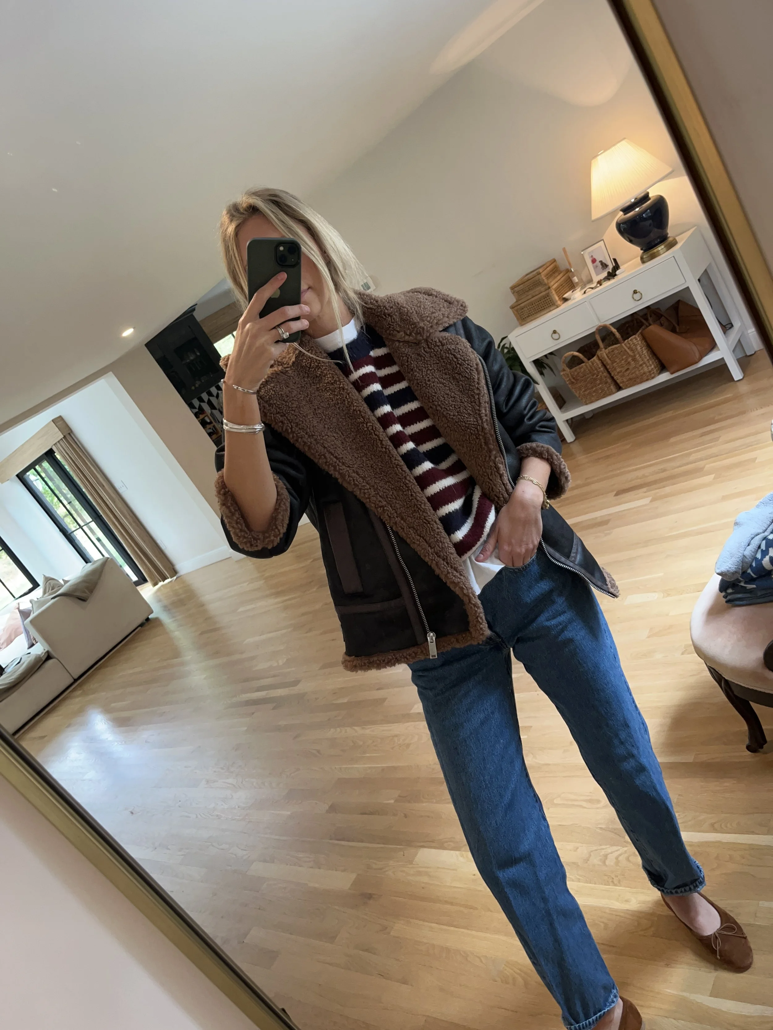 A woman taking a mirror selfie in a living room. She is wearing a striped sweater, a brown shearling-lined jacket, jeans, and brown loafers.