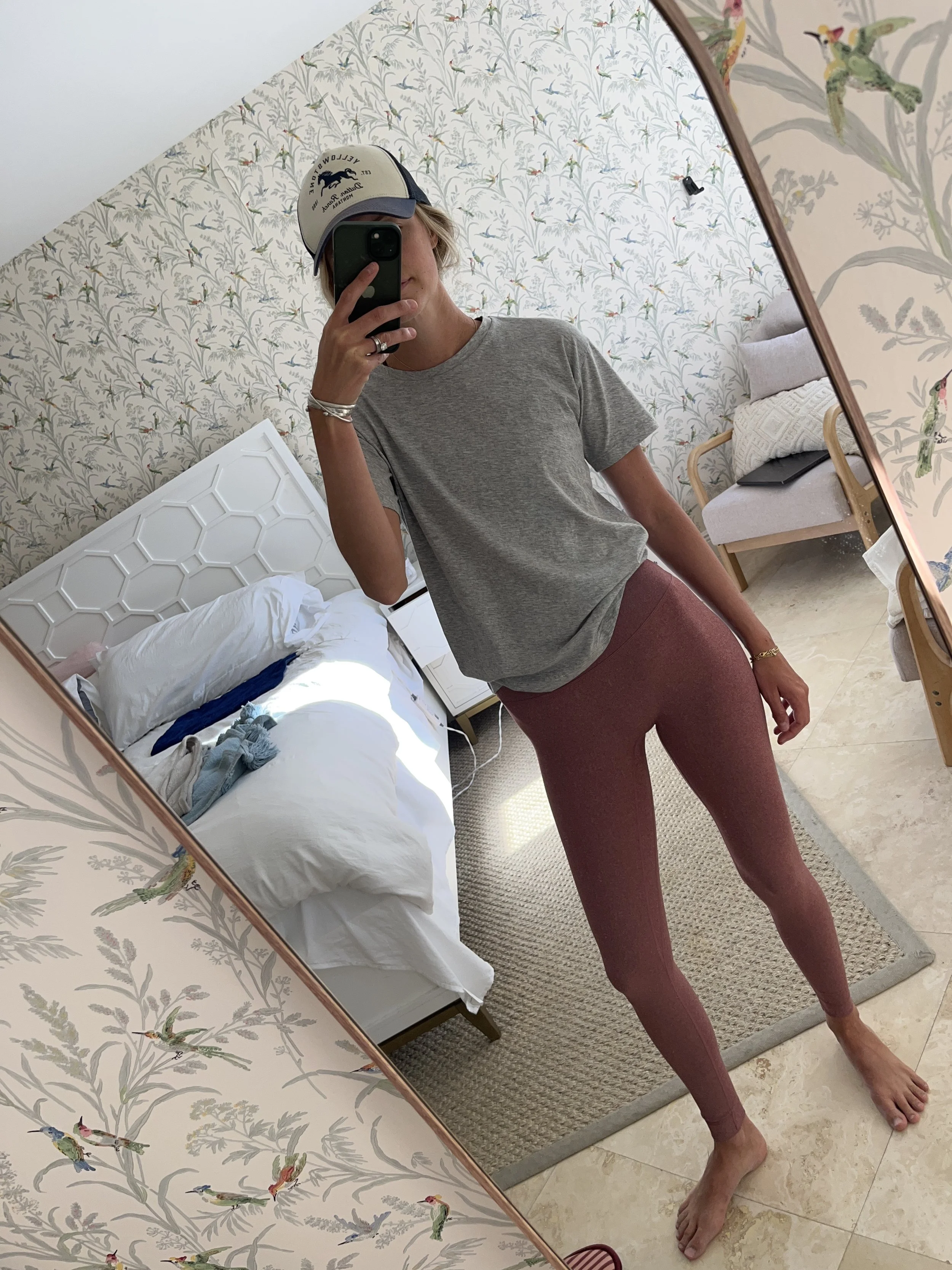 A woman taking a mirror selfie in a spacious, well-lit living room with hardwood floors, large windows, a white sofa, and a side table with a plant.