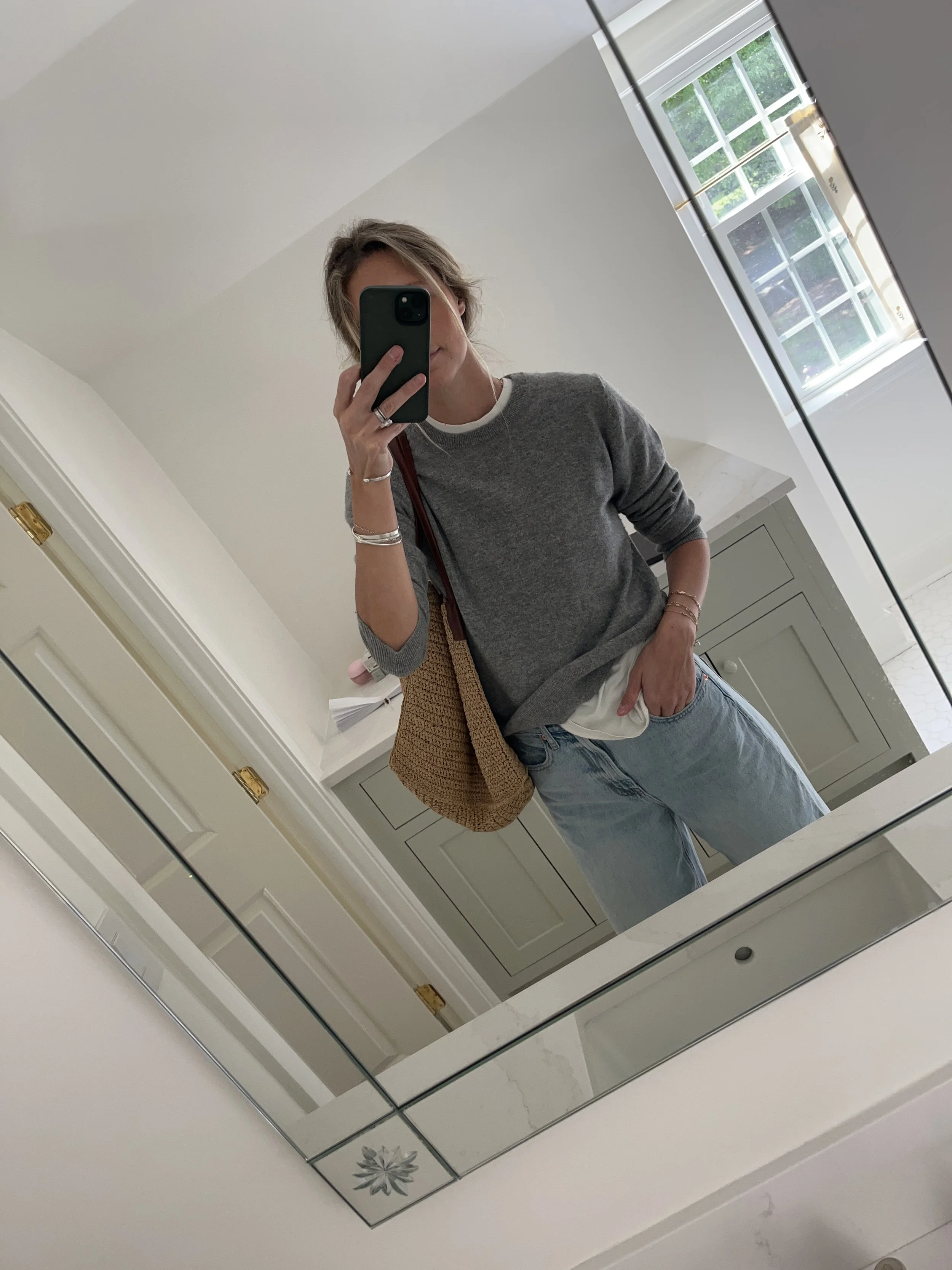 A woman taking a mirror selfie in a spacious living room with wooden floors, large windows, and minimal furniture, wearing a beige jacket, black top, blue jeans, and tan shoes.