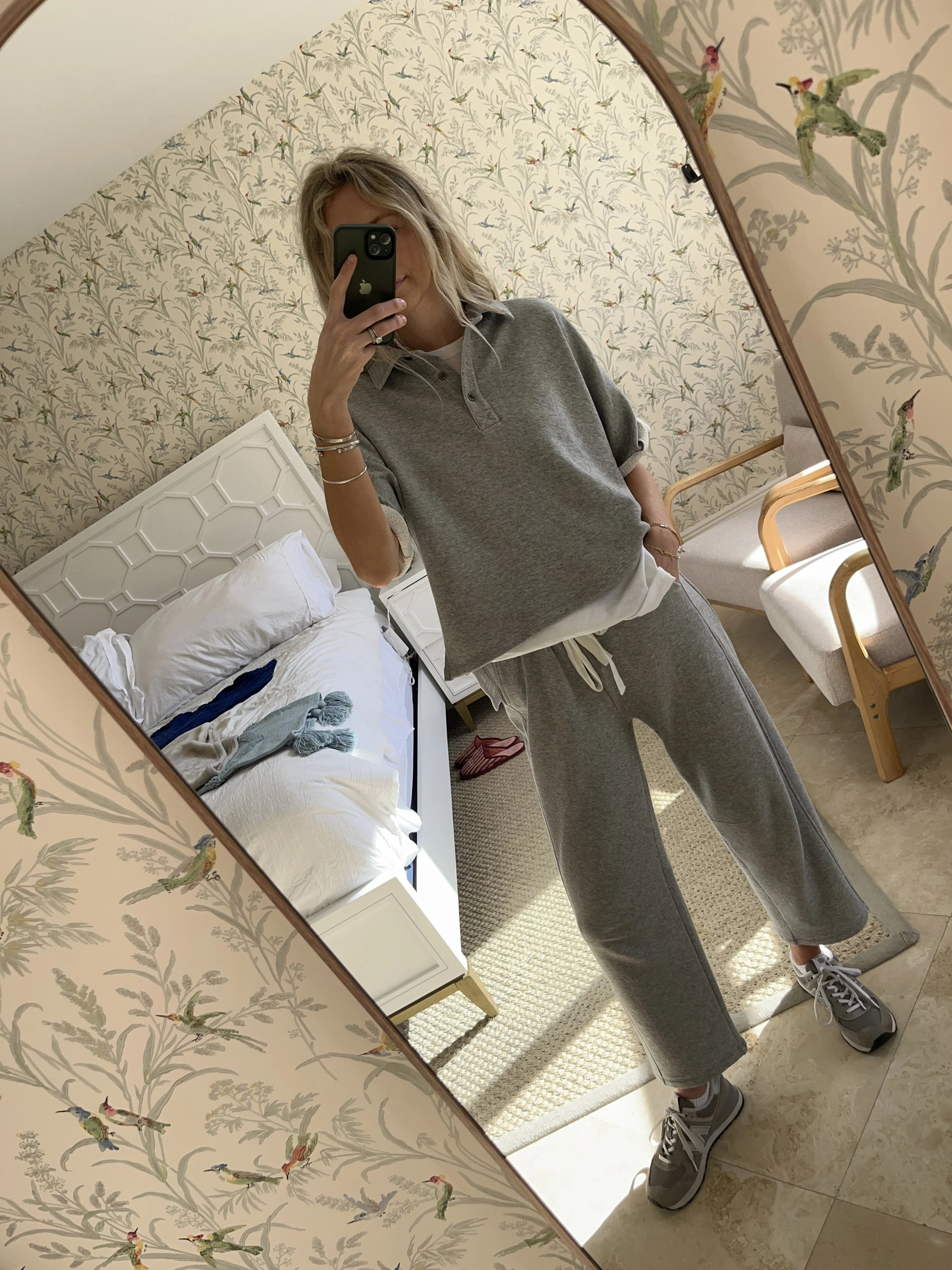 A woman taking a mirror selfie in a spacious living room with wooden floors, large windows, and minimal furniture, wearing a beige jacket, black top, blue jeans, and tan shoes.