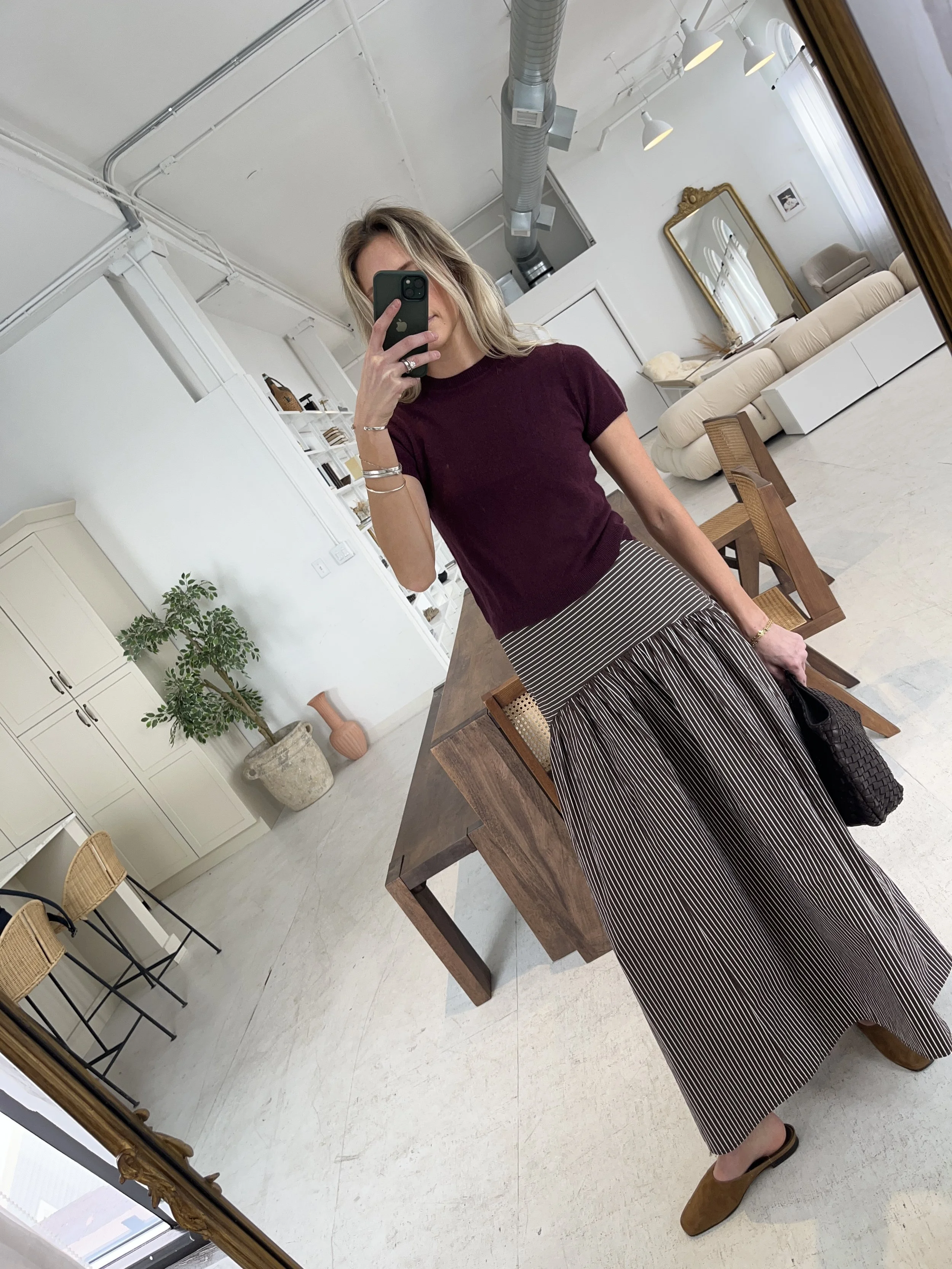 A woman taking a mirror selfie in a spacious living room with wooden floors, large windows, and minimal furniture, wearing a beige jacket, black top, blue jeans, and tan shoes.