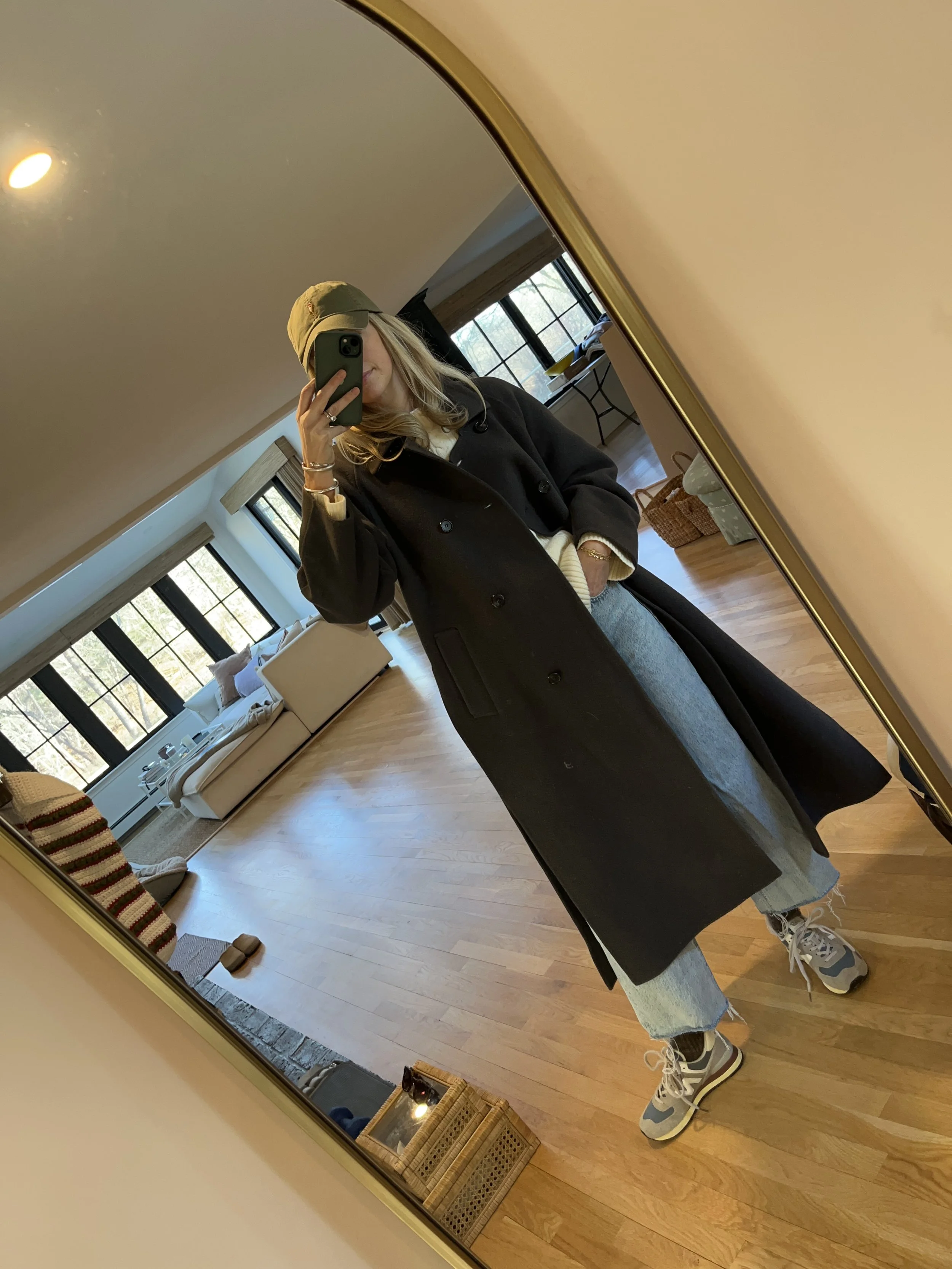 A woman taking a mirror selfie in a spacious living room with wooden floors, large windows, and minimal furniture, wearing a beige jacket, black top, blue jeans, and tan shoes.