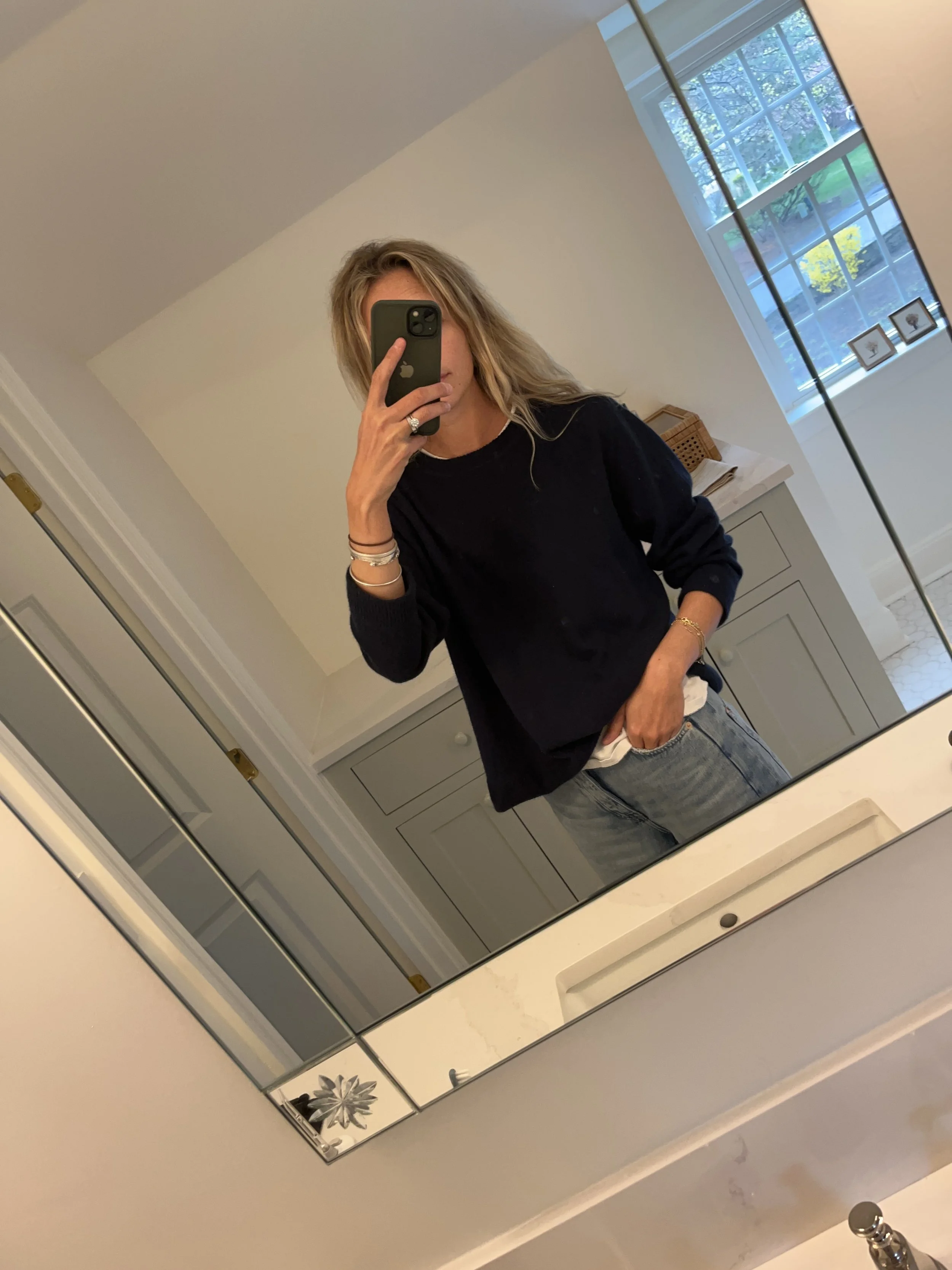 Person taking a mirror selfie in a room with wooden floors, wearing a striped brown and white long-sleeve shirt, light blue jeans, and black and white sneakers.