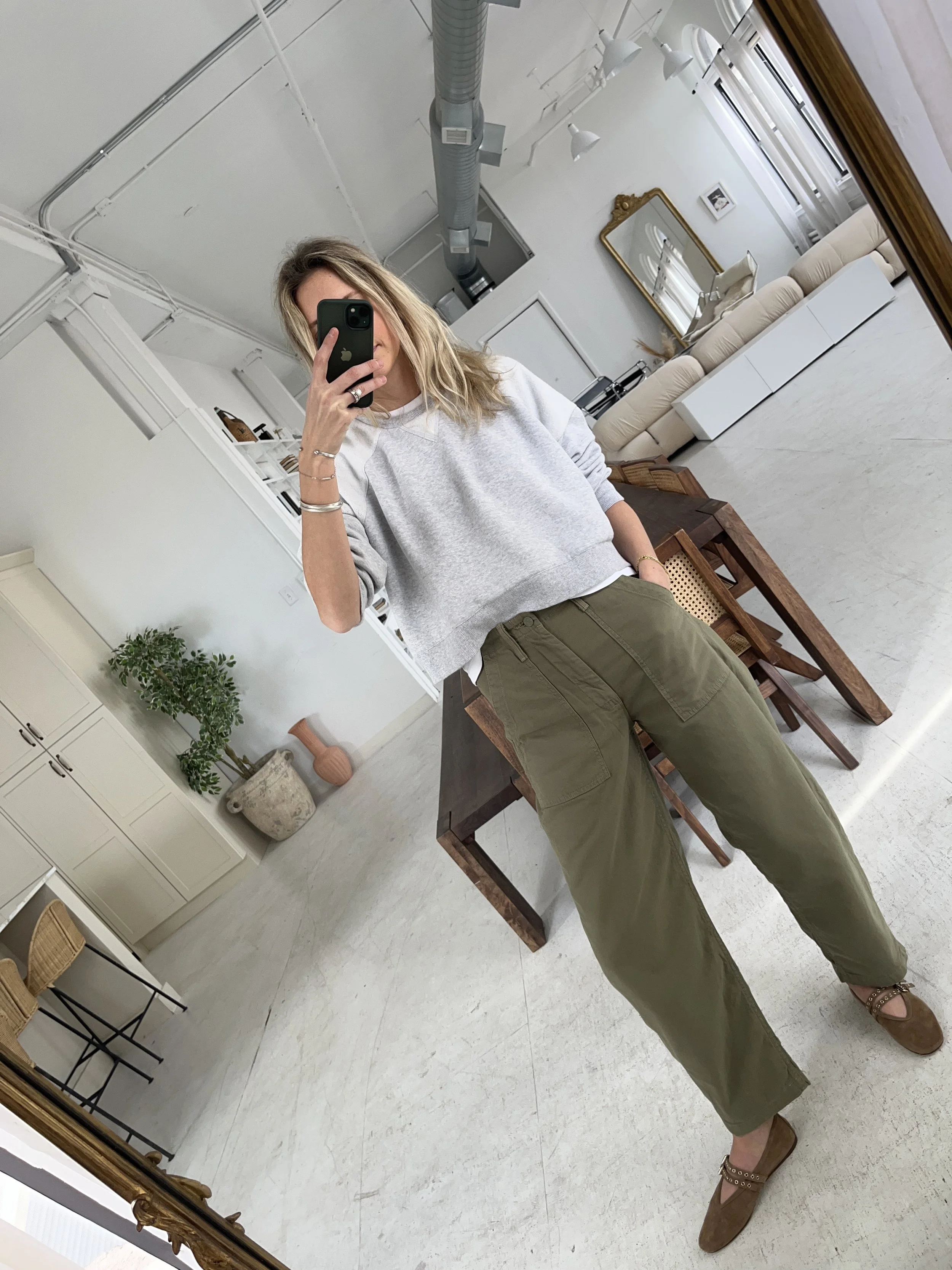 A woman taking a mirror selfie in a spacious living room with wooden floors, large windows, and minimal furniture, wearing a beige jacket, black top, blue jeans, and tan shoes.