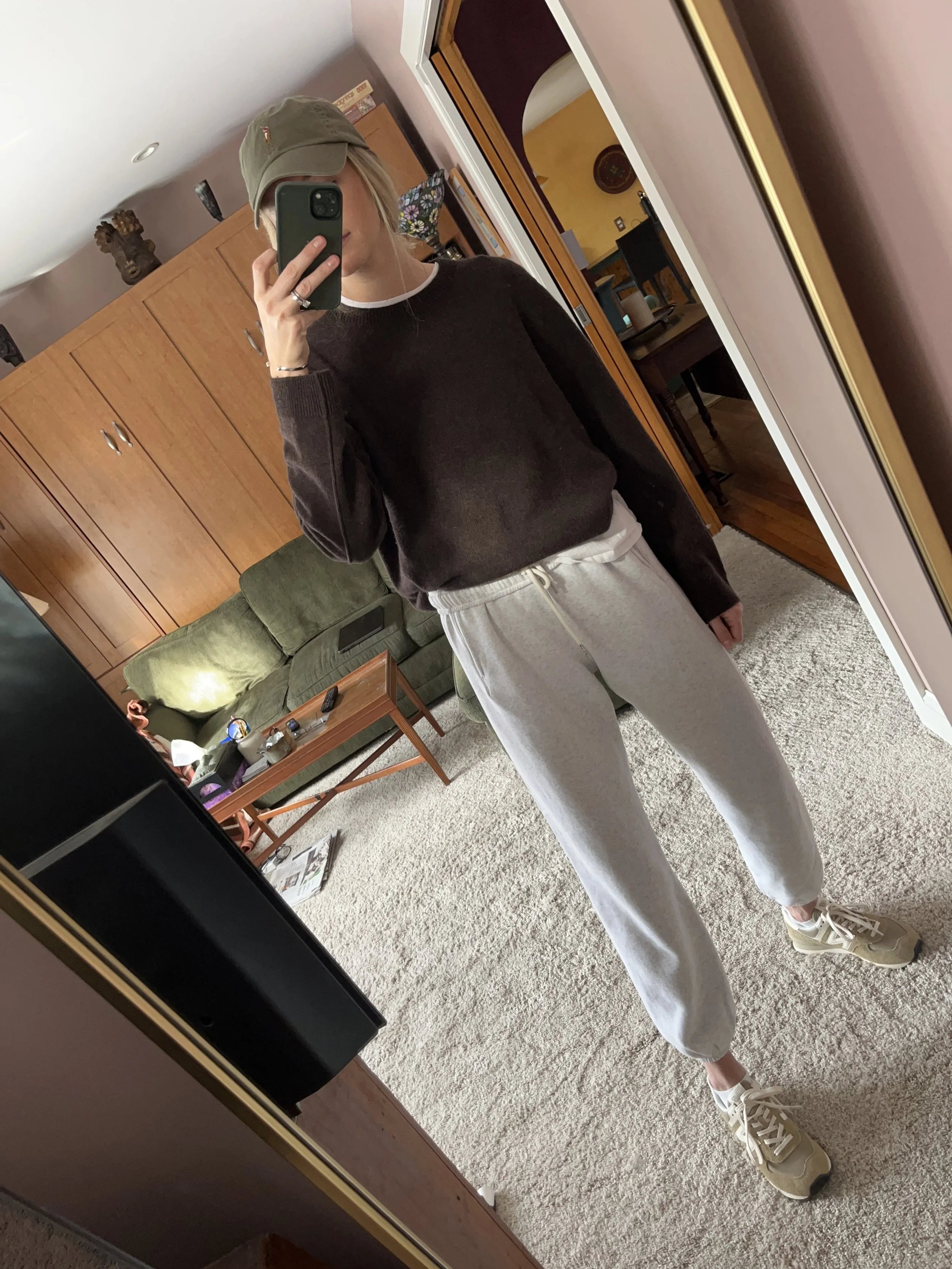 A woman taking a mirror selfie in a spacious, well-lit living room with hardwood floors, large windows, a white sofa, and a side table with a plant.