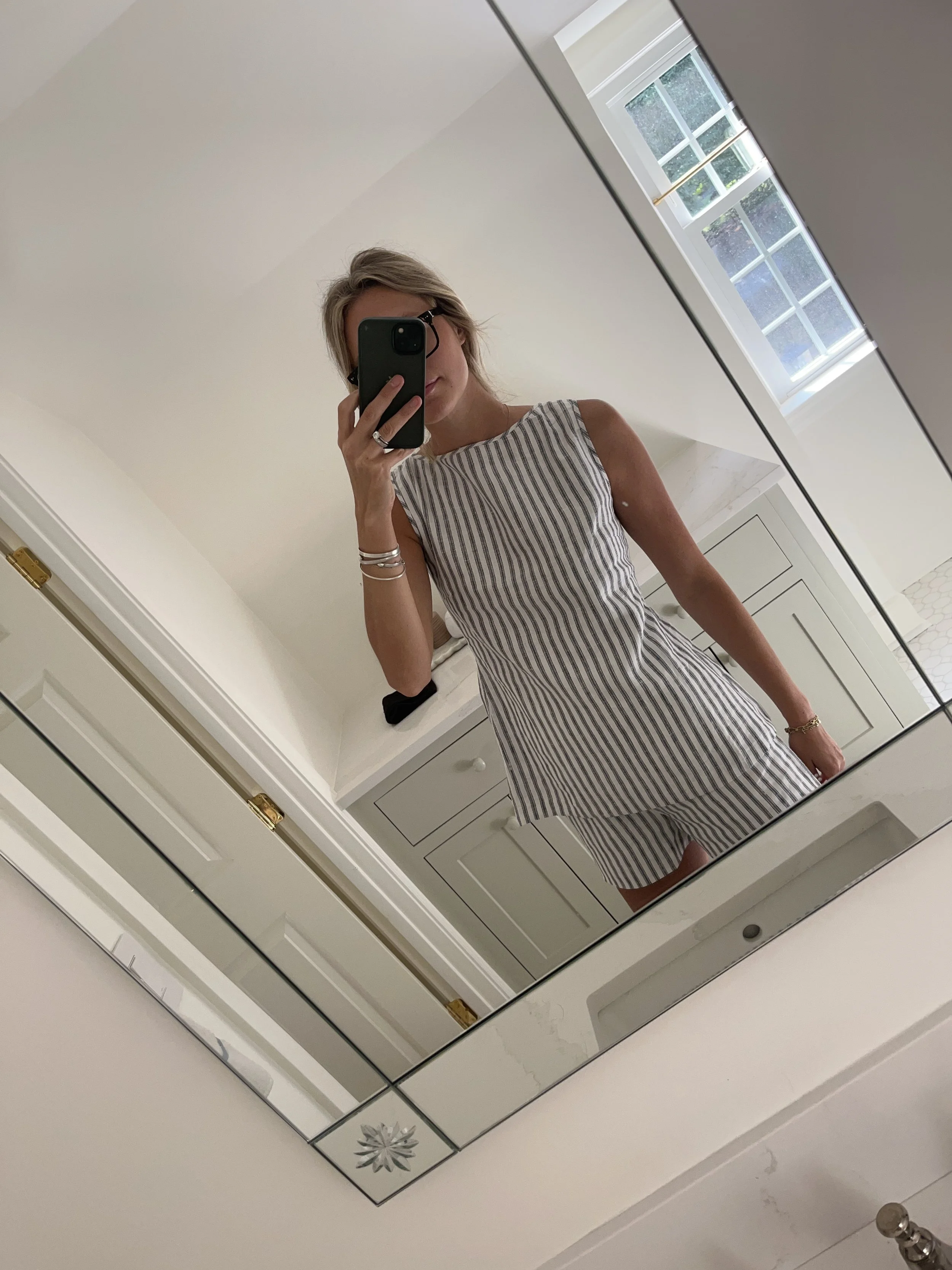 Person taking a mirror selfie in a room with wooden floors, wearing a striped brown and white long-sleeve shirt, light blue jeans, and black and white sneakers.