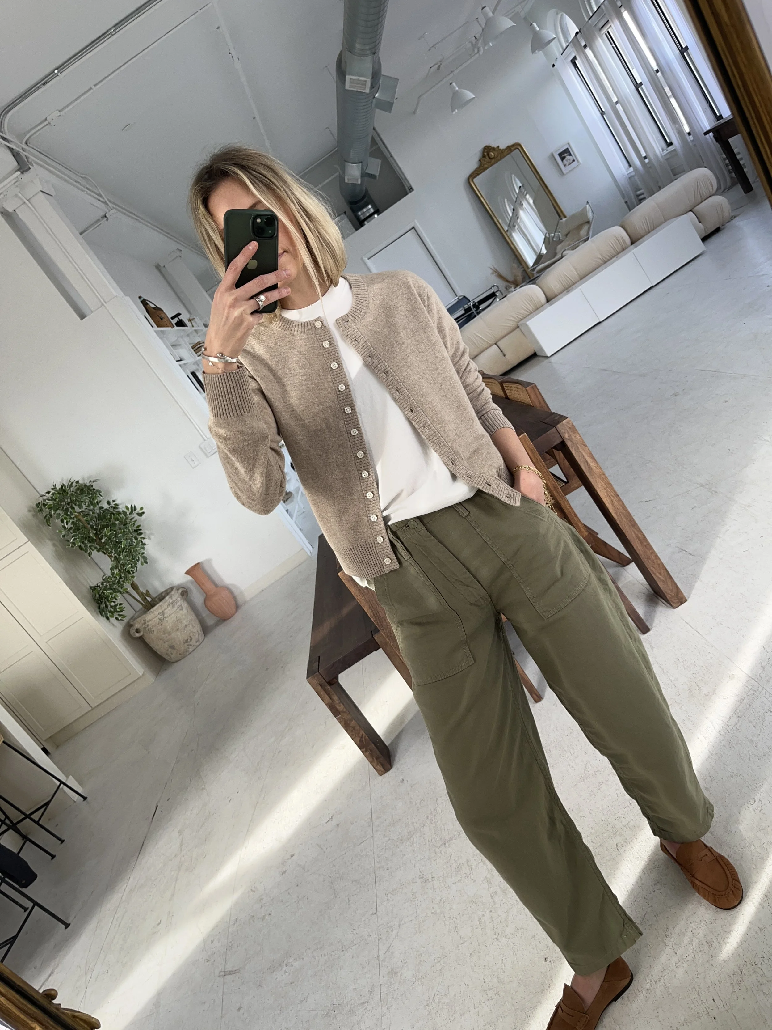 Person taking a mirror selfie in a room with wooden floors, wearing a striped brown and white long-sleeve shirt, light blue jeans, and black and white sneakers.