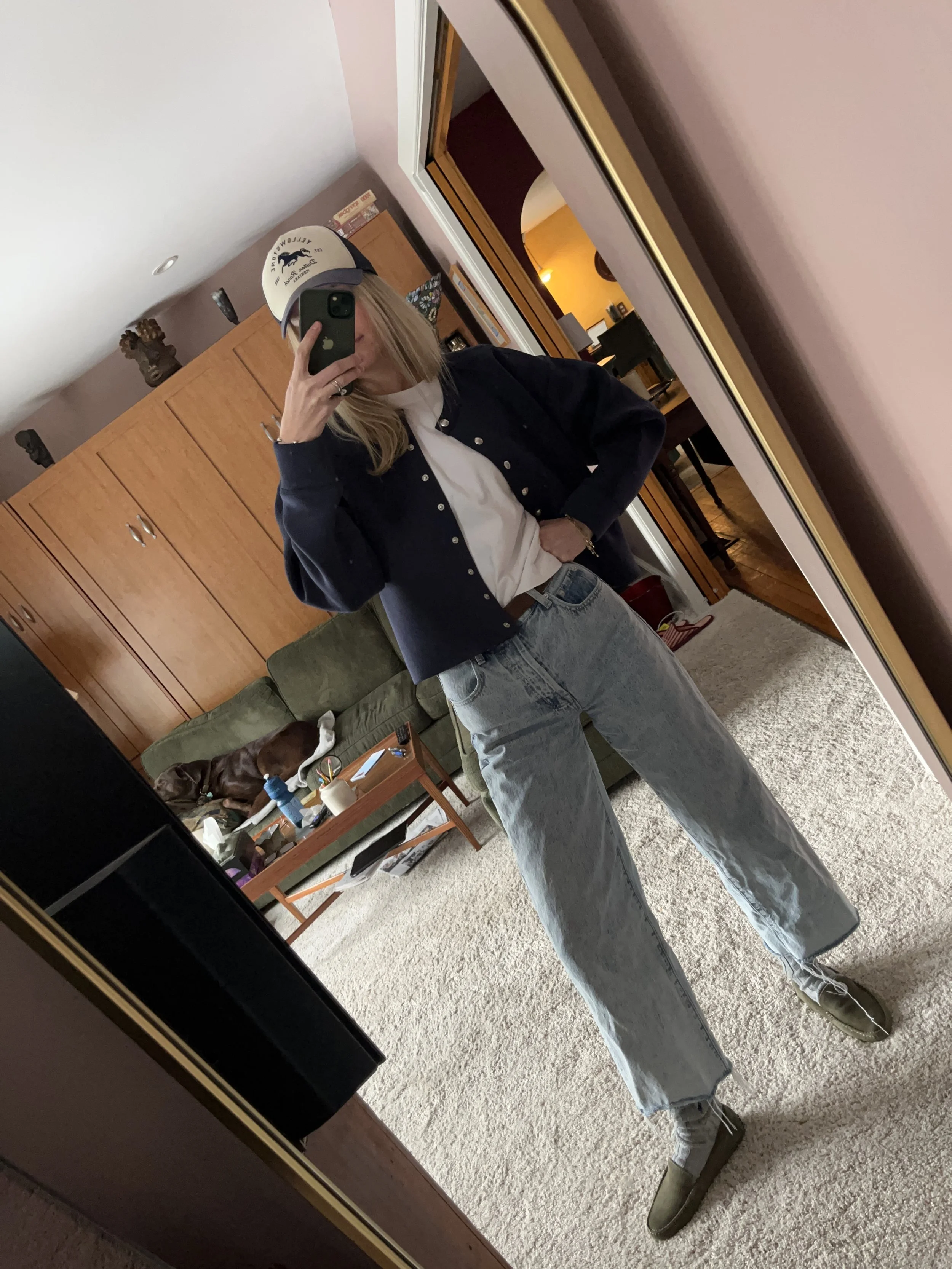 A woman taking a mirror selfie in a spacious living room with wooden floors, large windows, and minimal furniture, wearing a beige jacket, black top, blue jeans, and tan shoes.