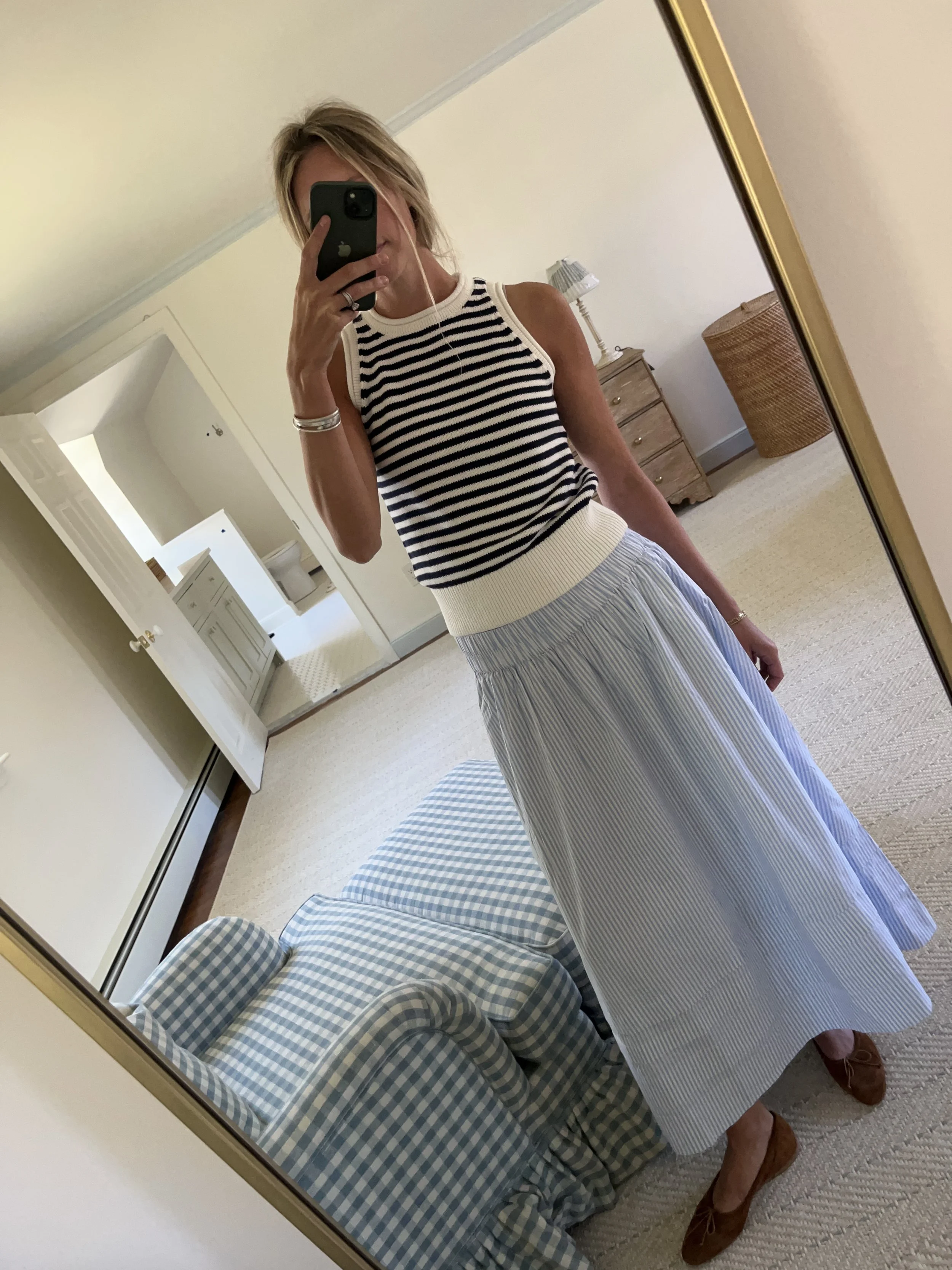 A woman taking a mirror selfie in a spacious living room with wooden floors, large windows, and minimal furniture, wearing a beige jacket, black top, blue jeans, and tan shoes.
