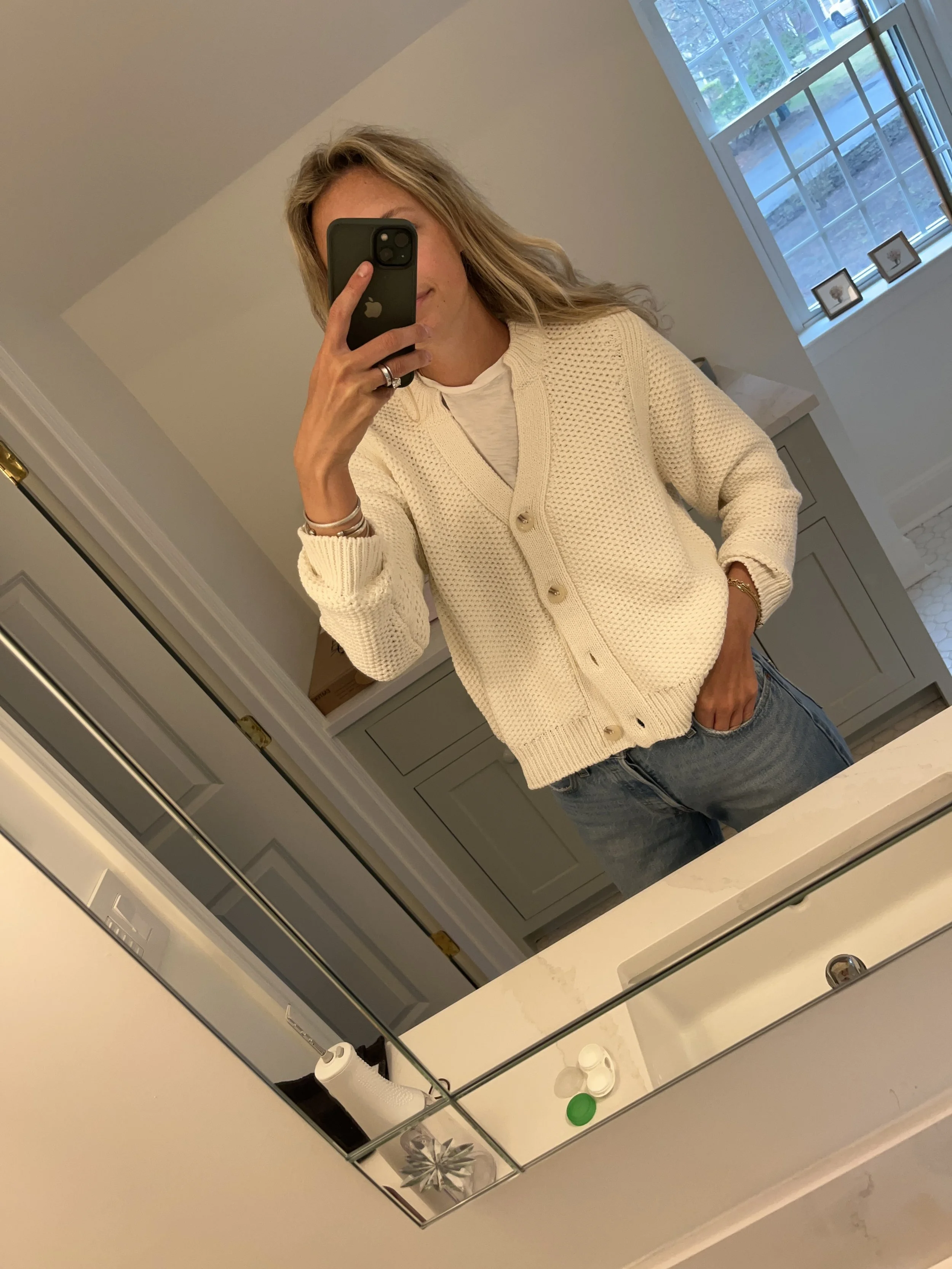A woman taking a mirror selfie in a spacious living room with wooden floors, large windows, and minimal furniture, wearing a beige jacket, black top, blue jeans, and tan shoes.