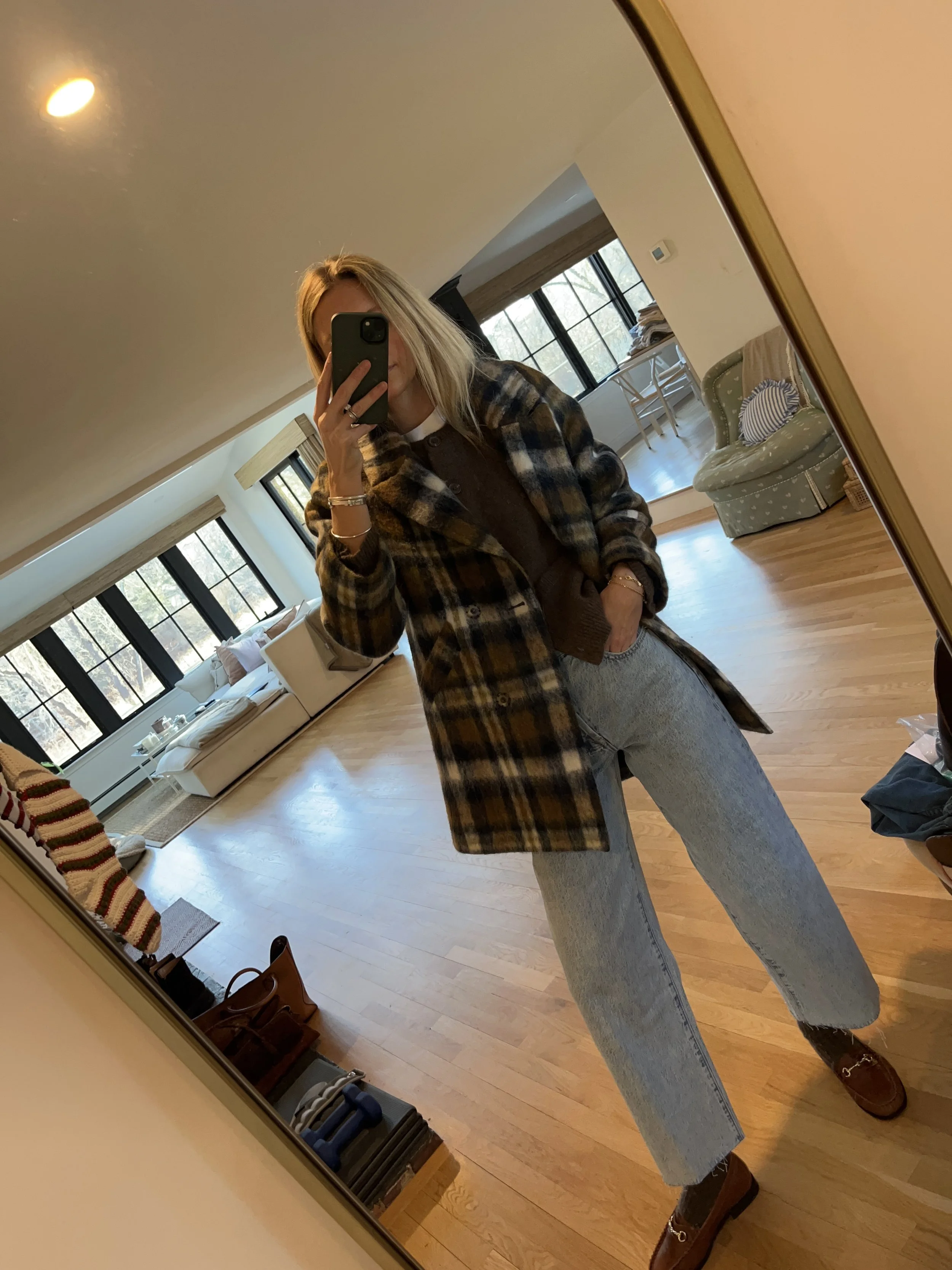 A woman taking a mirror selfie in a spacious, well-lit living room with hardwood floors, large windows, a white sofa, and a side table with a plant.