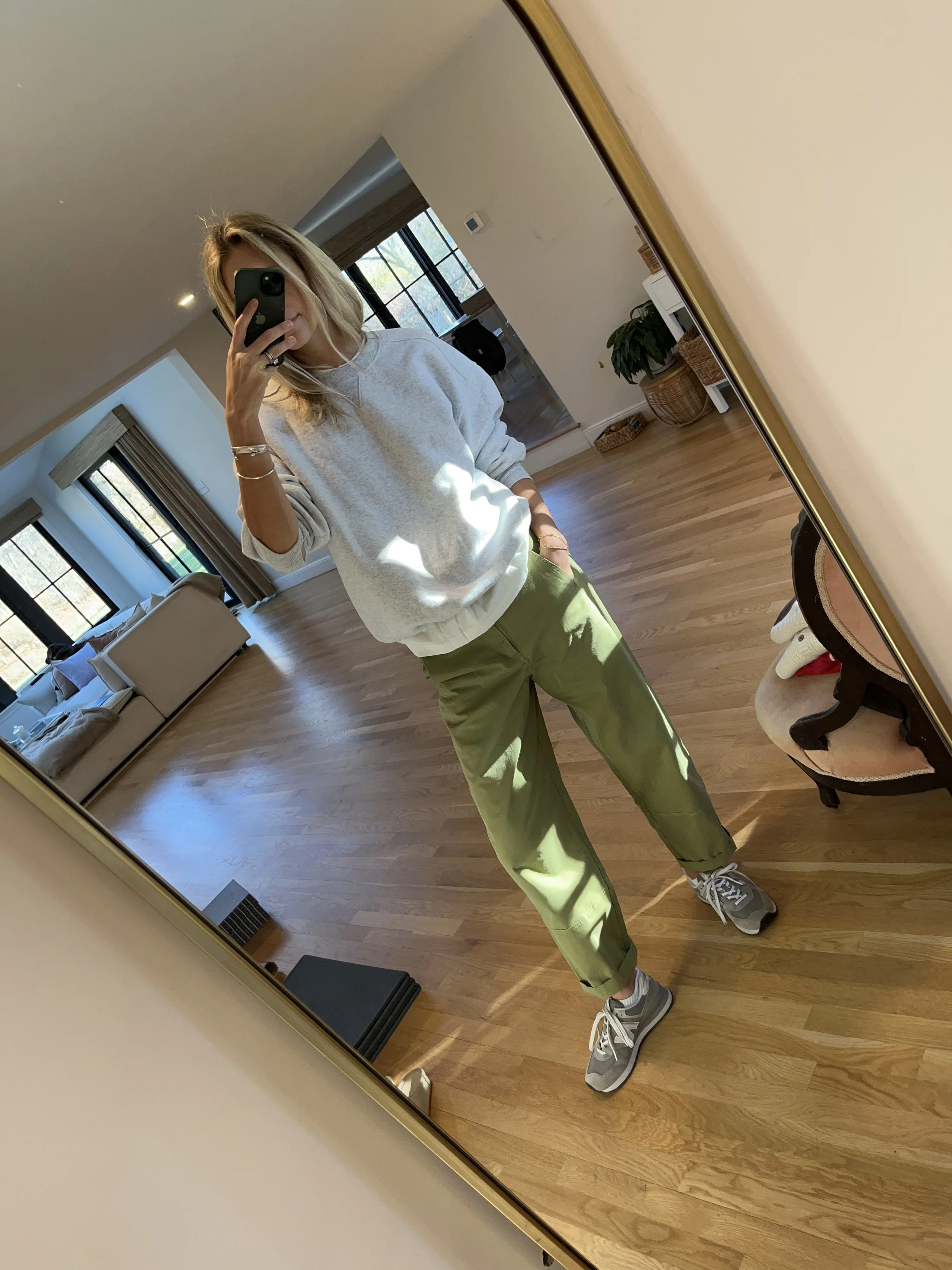 Person taking a mirror selfie in a room with wooden floors, wearing a striped brown and white long-sleeve shirt, light blue jeans, and black and white sneakers.