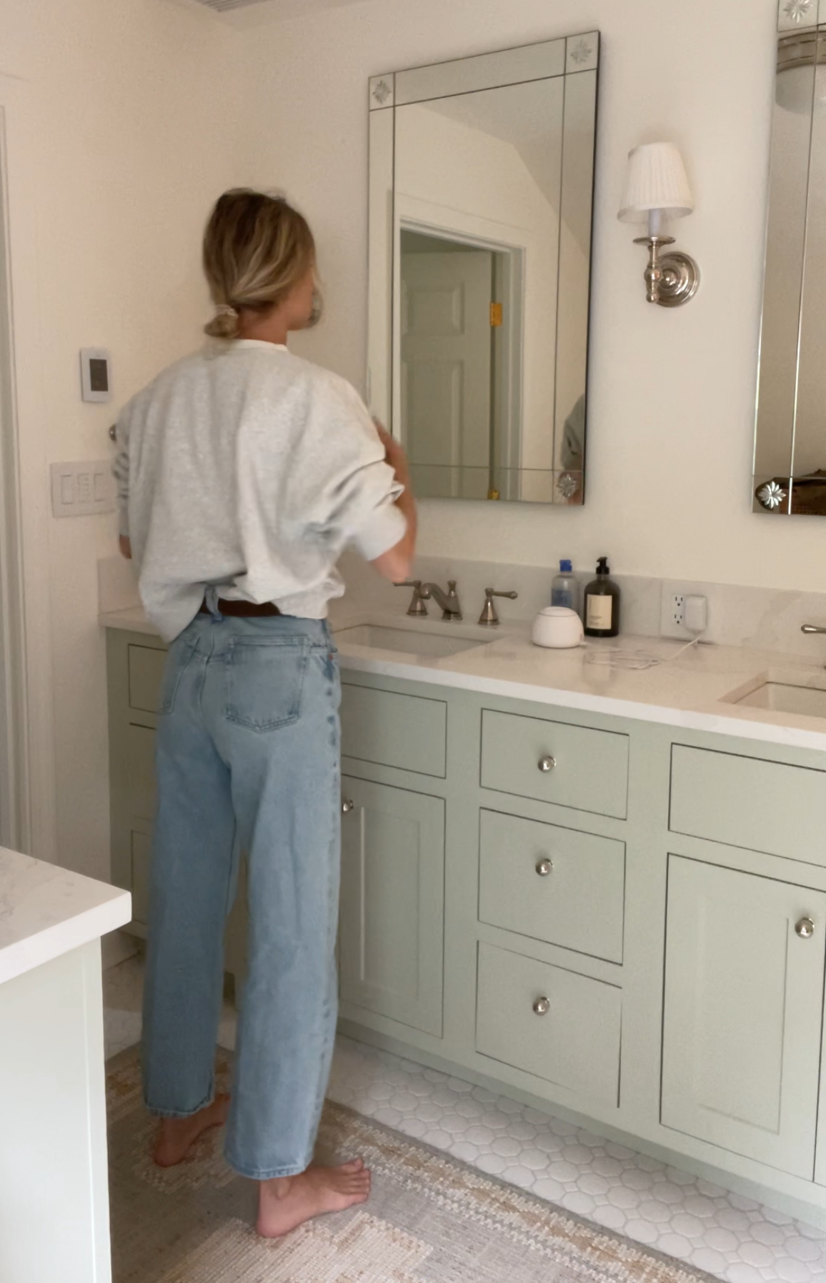 A woman taking a mirror selfie in a spacious living room with wooden floors, large windows, and minimal furniture, wearing a beige jacket, black top, blue jeans, and tan shoes.