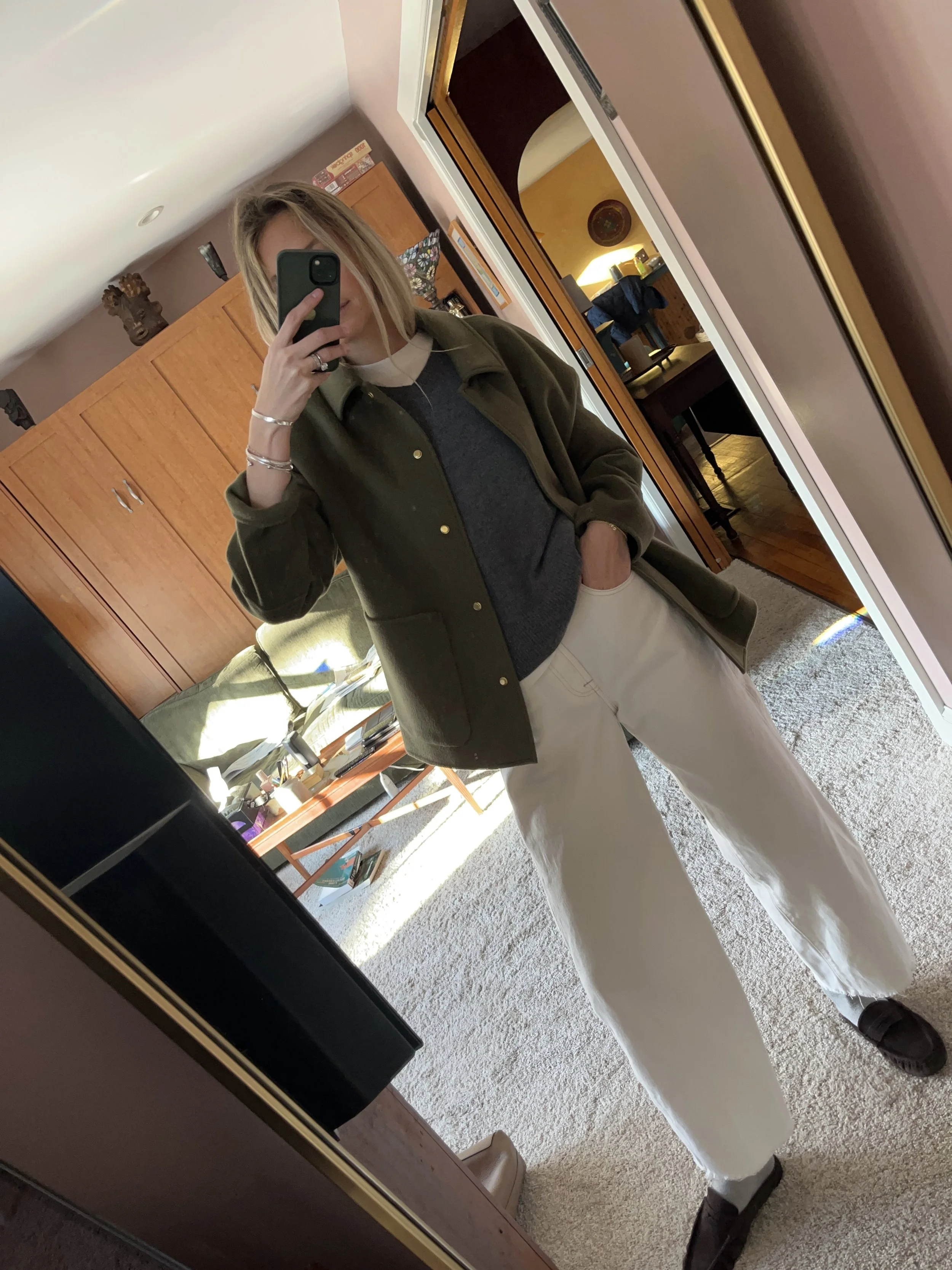 Person taking a mirror selfie in a room with wooden floors, wearing a striped brown and white long-sleeve shirt, light blue jeans, and black and white sneakers.