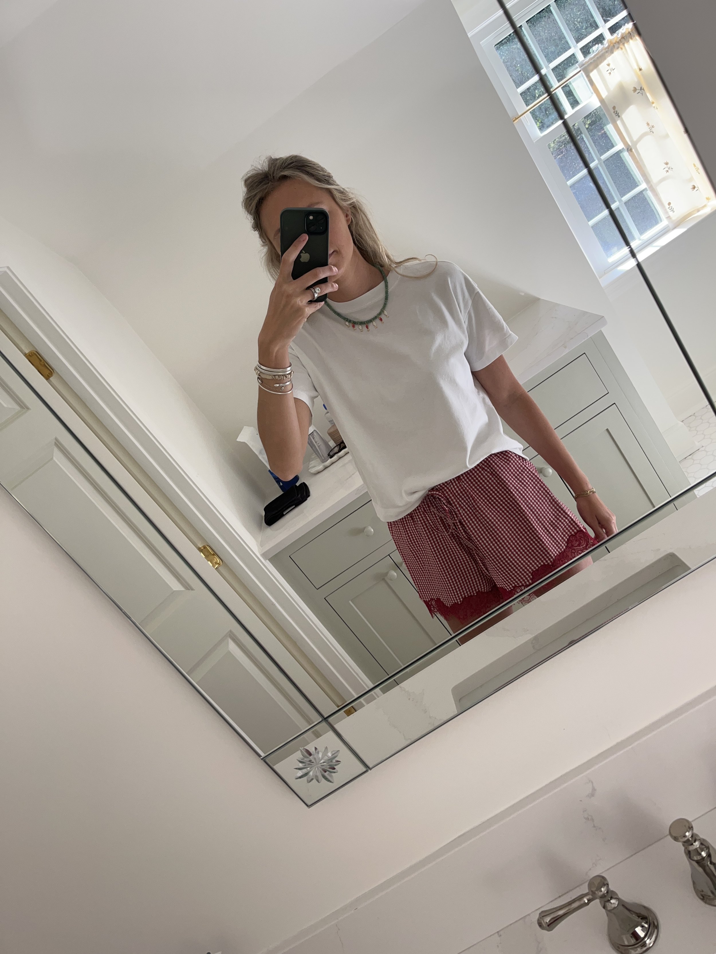 A woman taking a mirror selfie in a spacious living room with wooden floors, large windows, and minimal furniture, wearing a beige jacket, black top, blue jeans, and tan shoes.