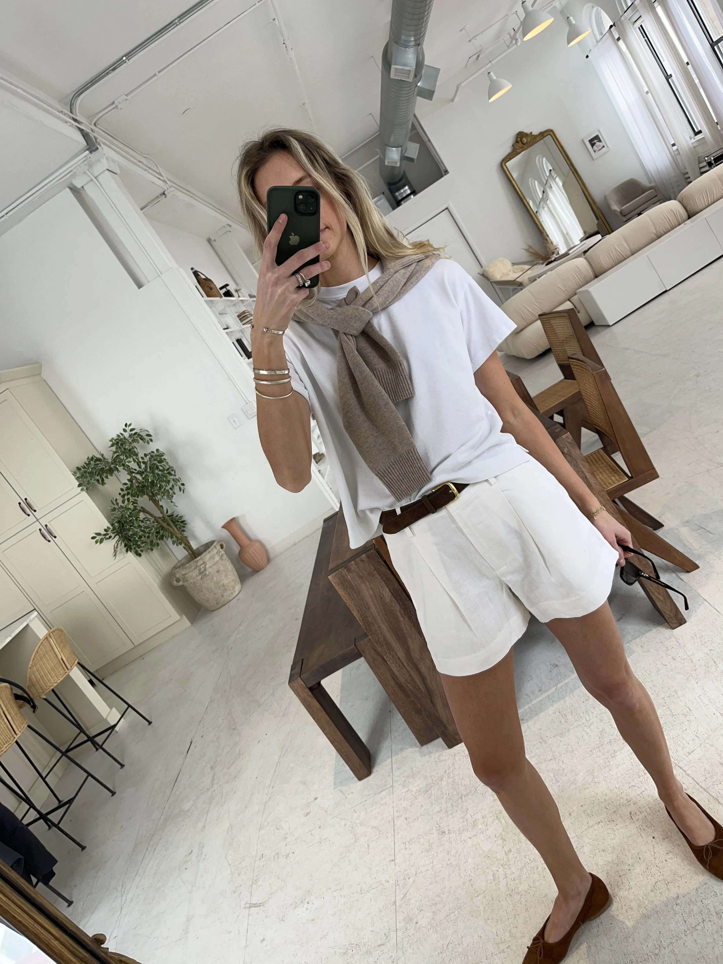 A woman taking a mirror selfie in a spacious, well-lit living room with hardwood floors, large windows, a white sofa, and a side table with a plant.