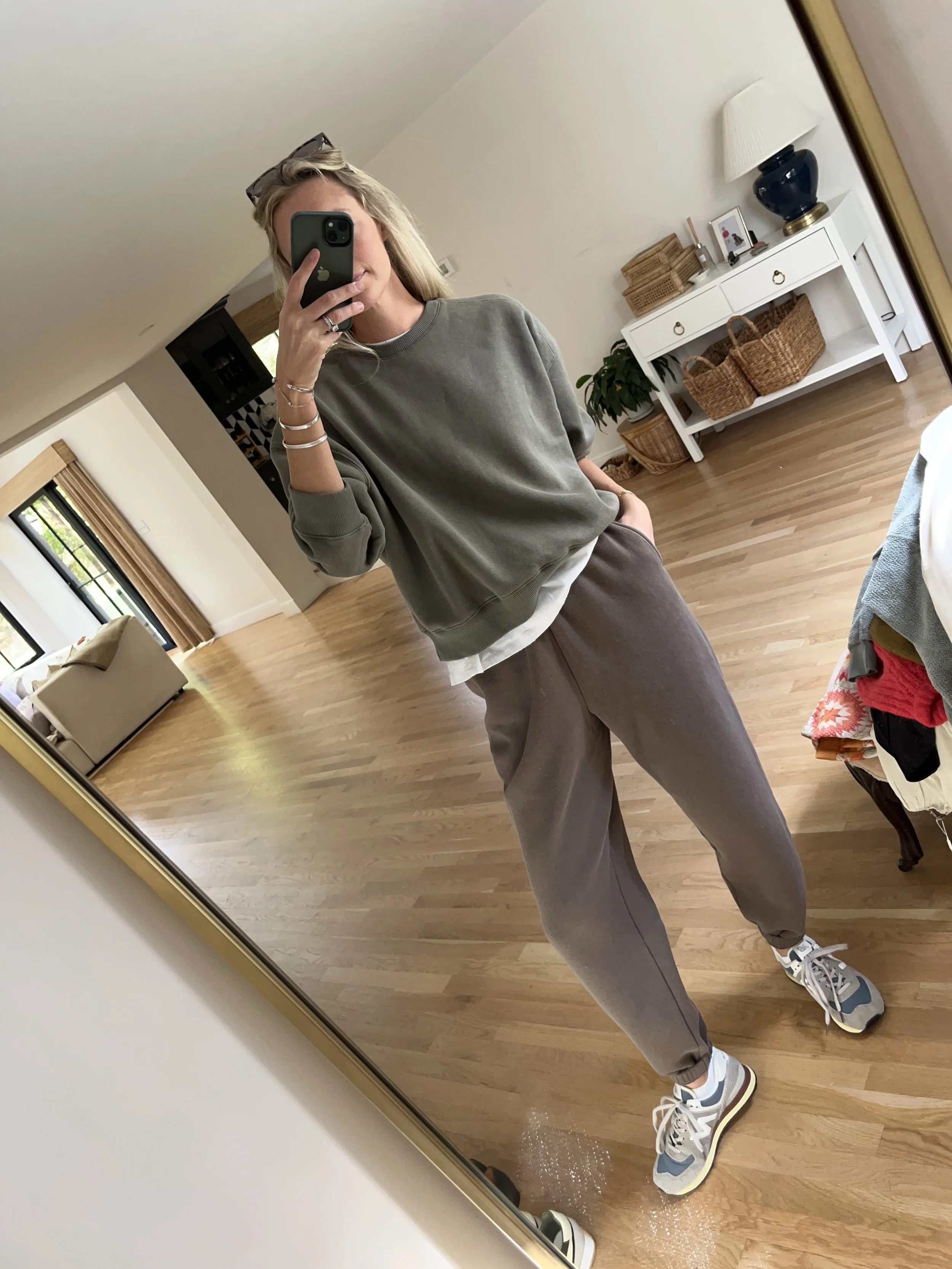 Woman taking a mirror selfie in a cozy living room, wearing a gray sweatshirt, beige joggers, and sneakers, with casual accessories.