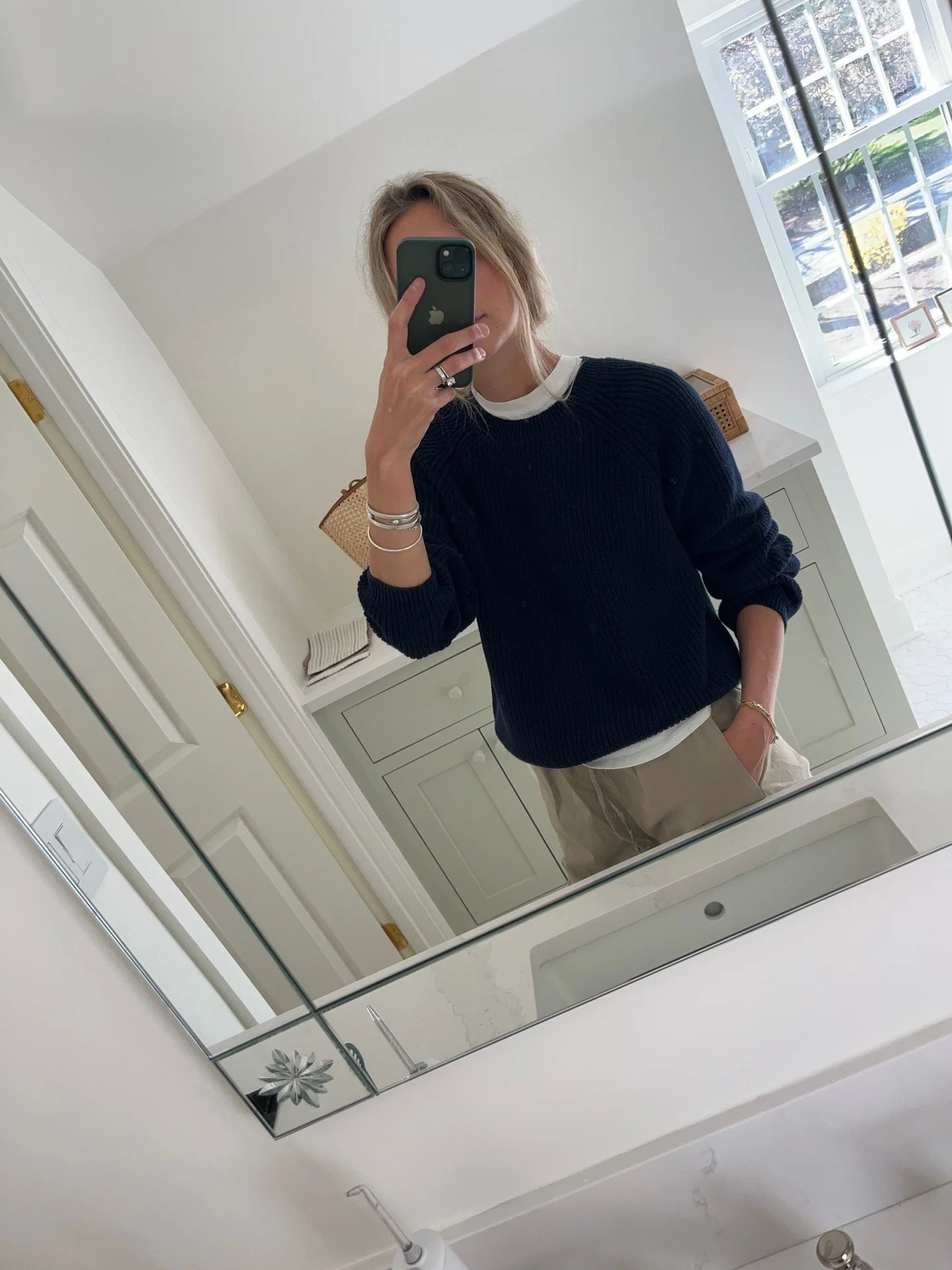 A woman taking a mirror selfie in a spacious living room with wooden floors, large windows, and minimal furniture, wearing a beige jacket, black top, blue jeans, and tan shoes.