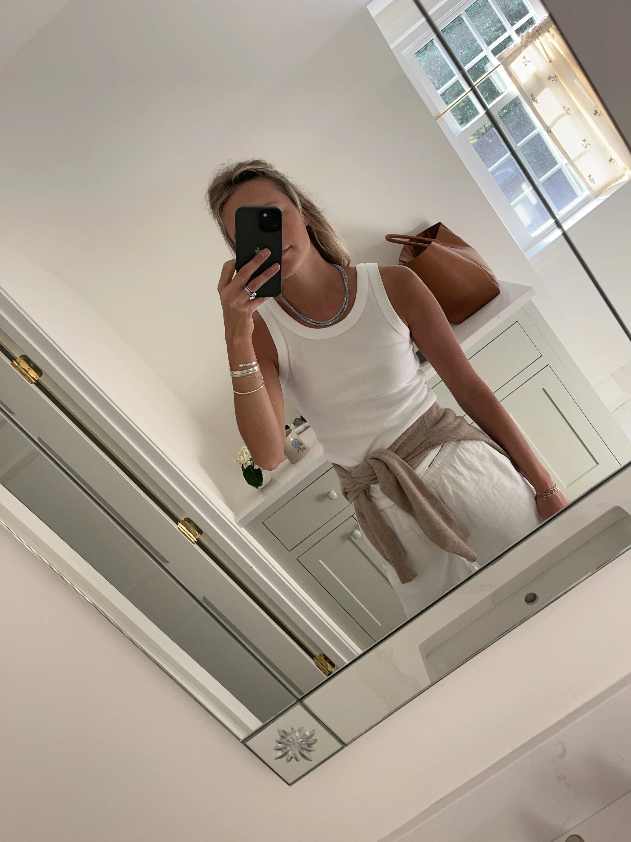 Person taking a mirror selfie in a room with wooden floors, wearing a striped brown and white long-sleeve shirt, light blue jeans, and black and white sneakers.