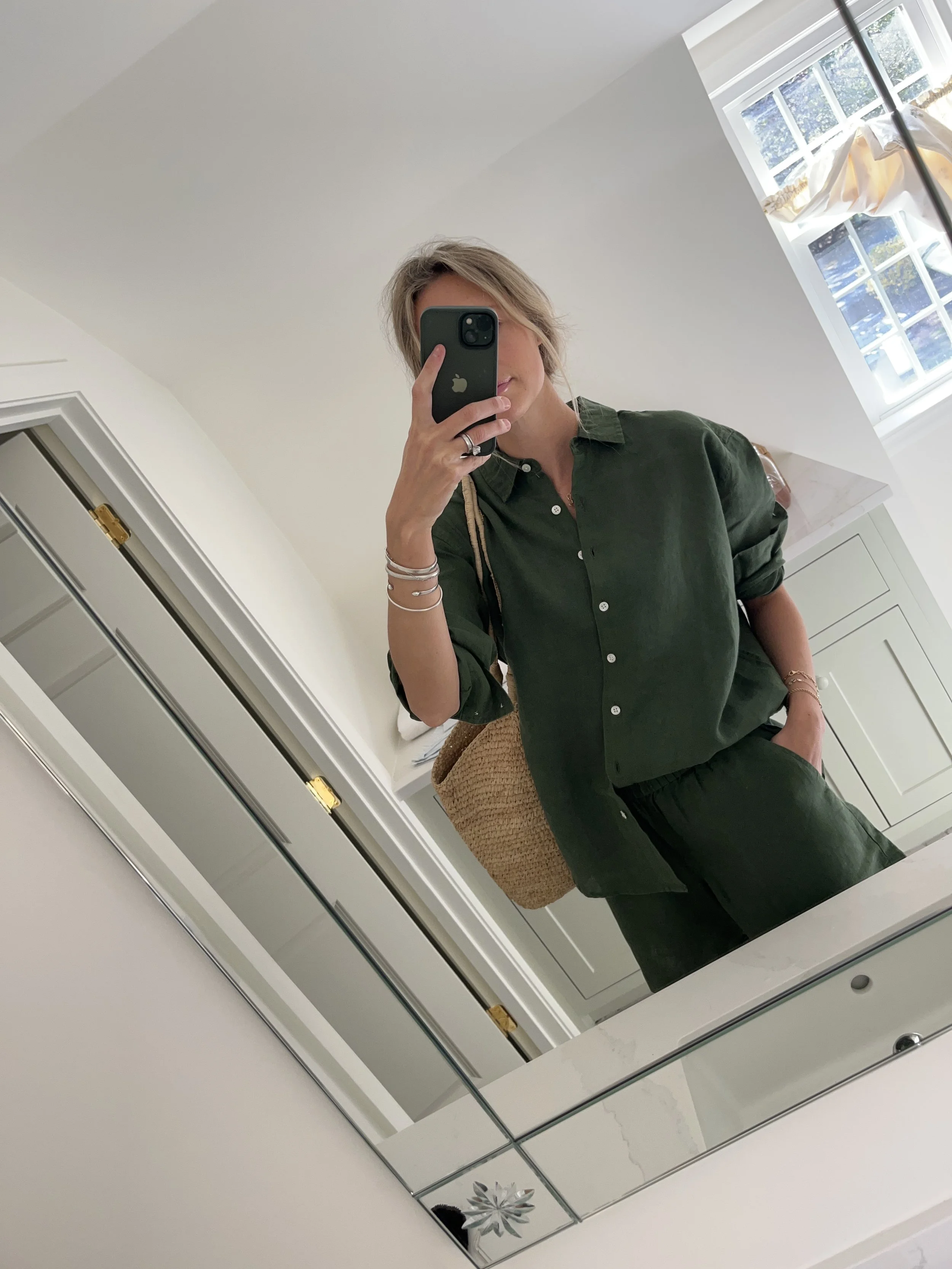 A woman taking a mirror selfie in a spacious, well-lit living room with hardwood floors, large windows, a white sofa, and a side table with a plant.