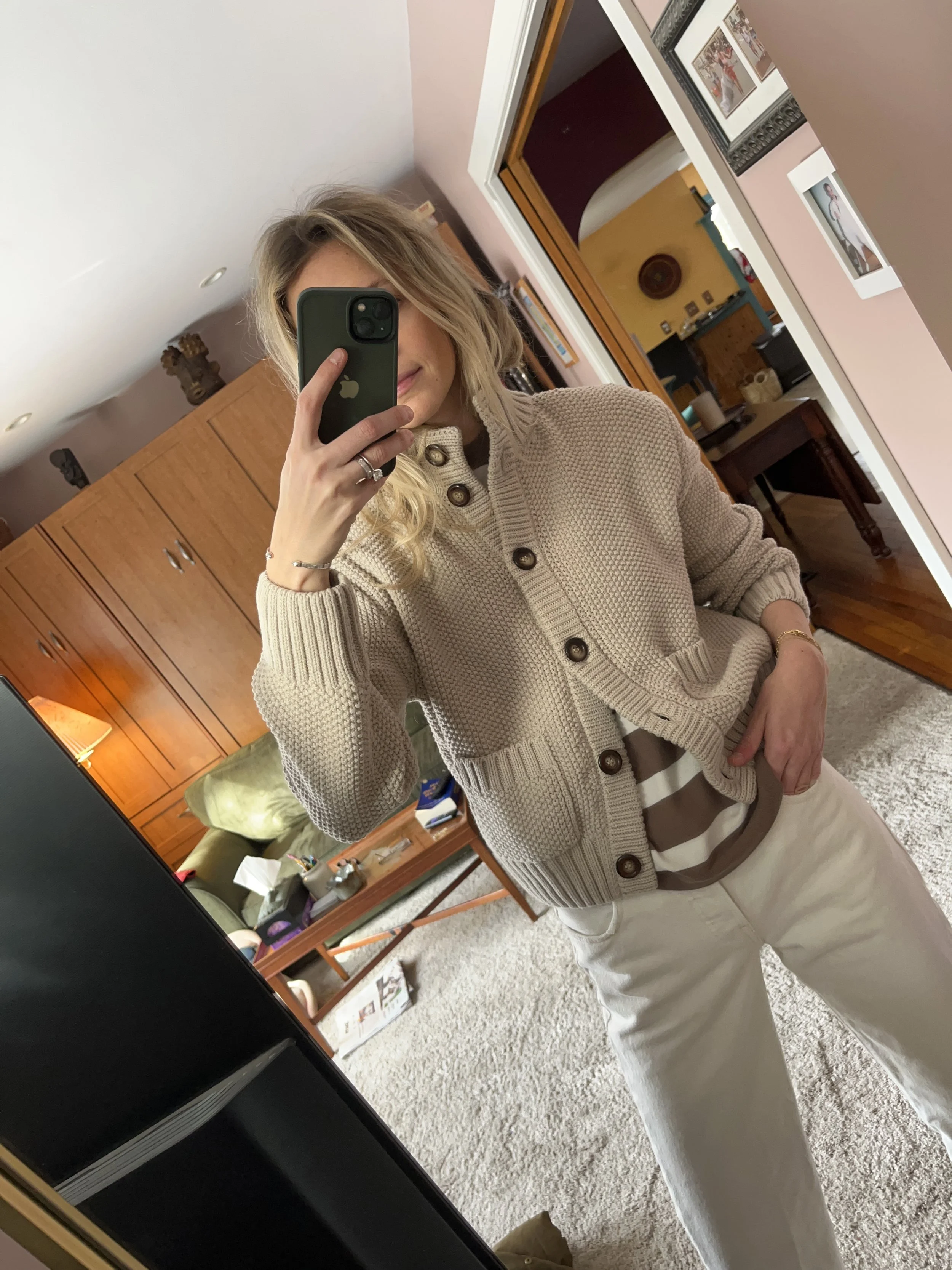 A woman taking a mirror selfie in a spacious living room with wooden floors, large windows, and minimal furniture, wearing a beige jacket, black top, blue jeans, and tan shoes.