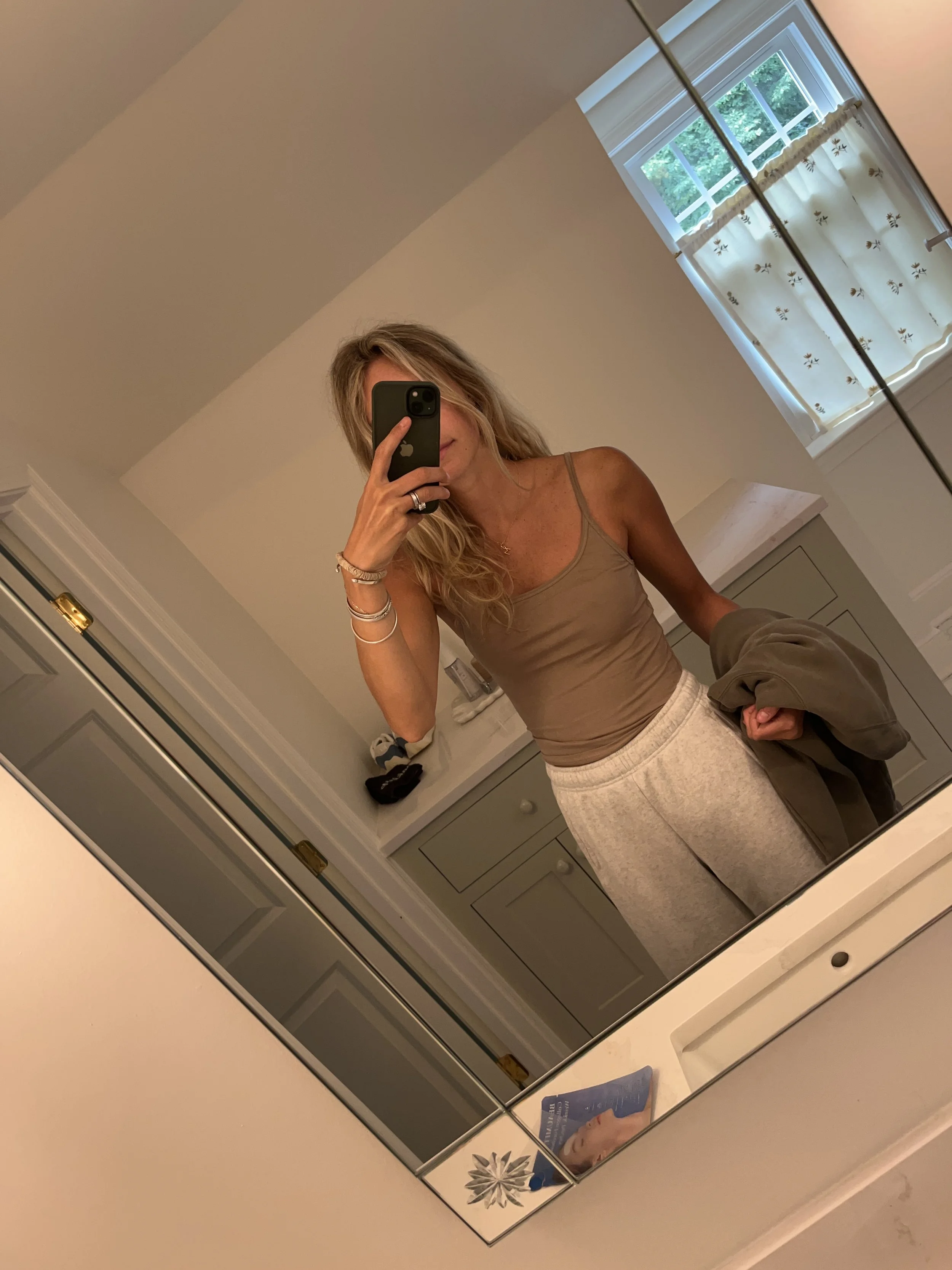 A woman taking a mirror selfie in a spacious, well-lit living room with hardwood floors, large windows, a white sofa, and a side table with a plant.
