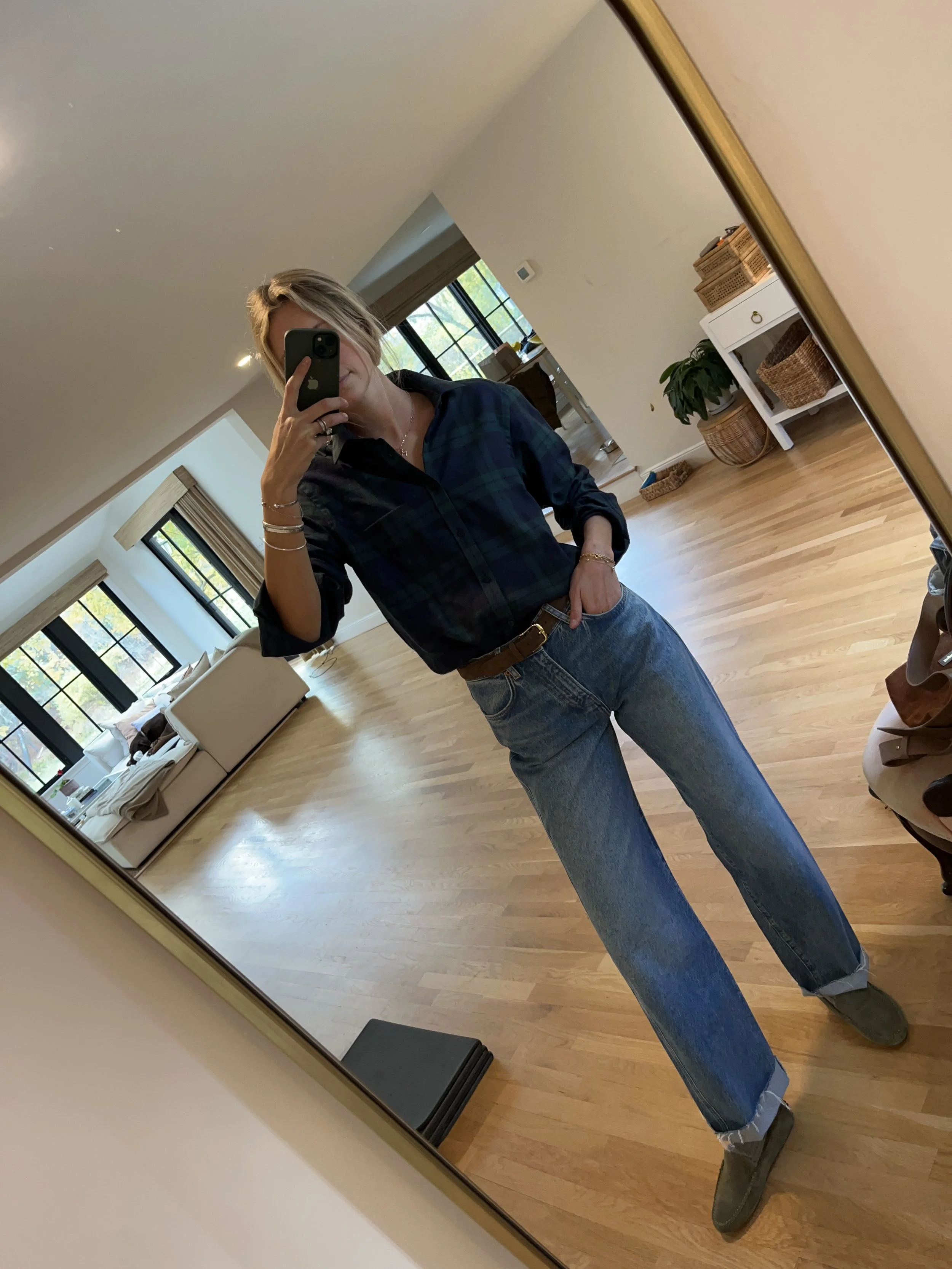 A woman taking a mirror selfie in a spacious, well-lit living room with hardwood floors, large windows, a white sofa, and a side table with a plant.