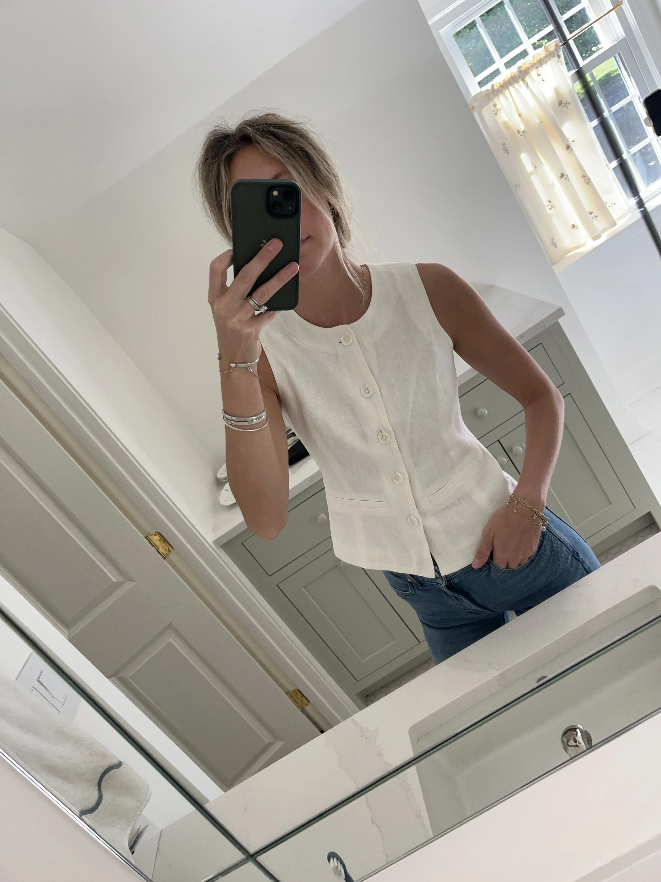 A woman taking a mirror selfie in a spacious, well-lit living room with hardwood floors, large windows, a white sofa, and a side table with a plant.