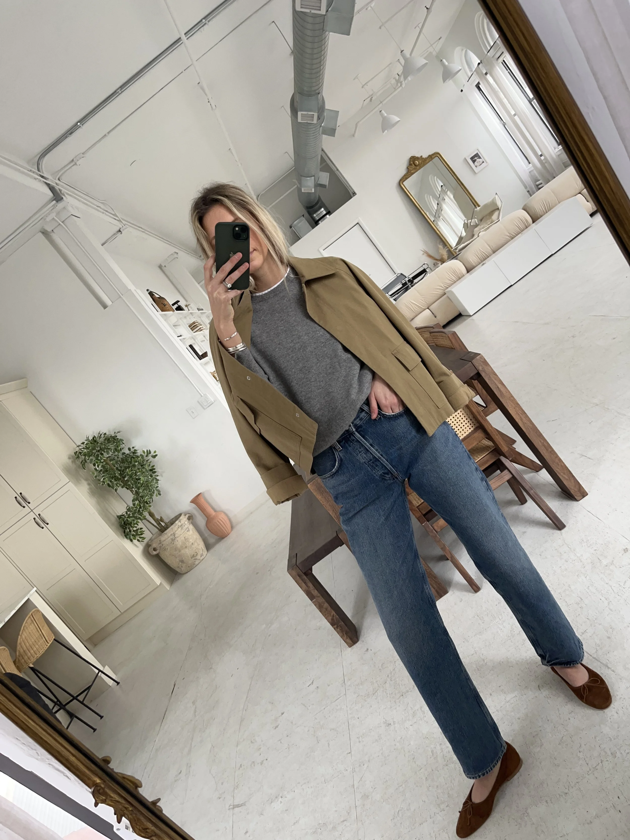 A woman taking a mirror selfie in a spacious living room with wooden floors, large windows, and minimal furniture, wearing a beige jacket, black top, blue jeans, and tan shoes.