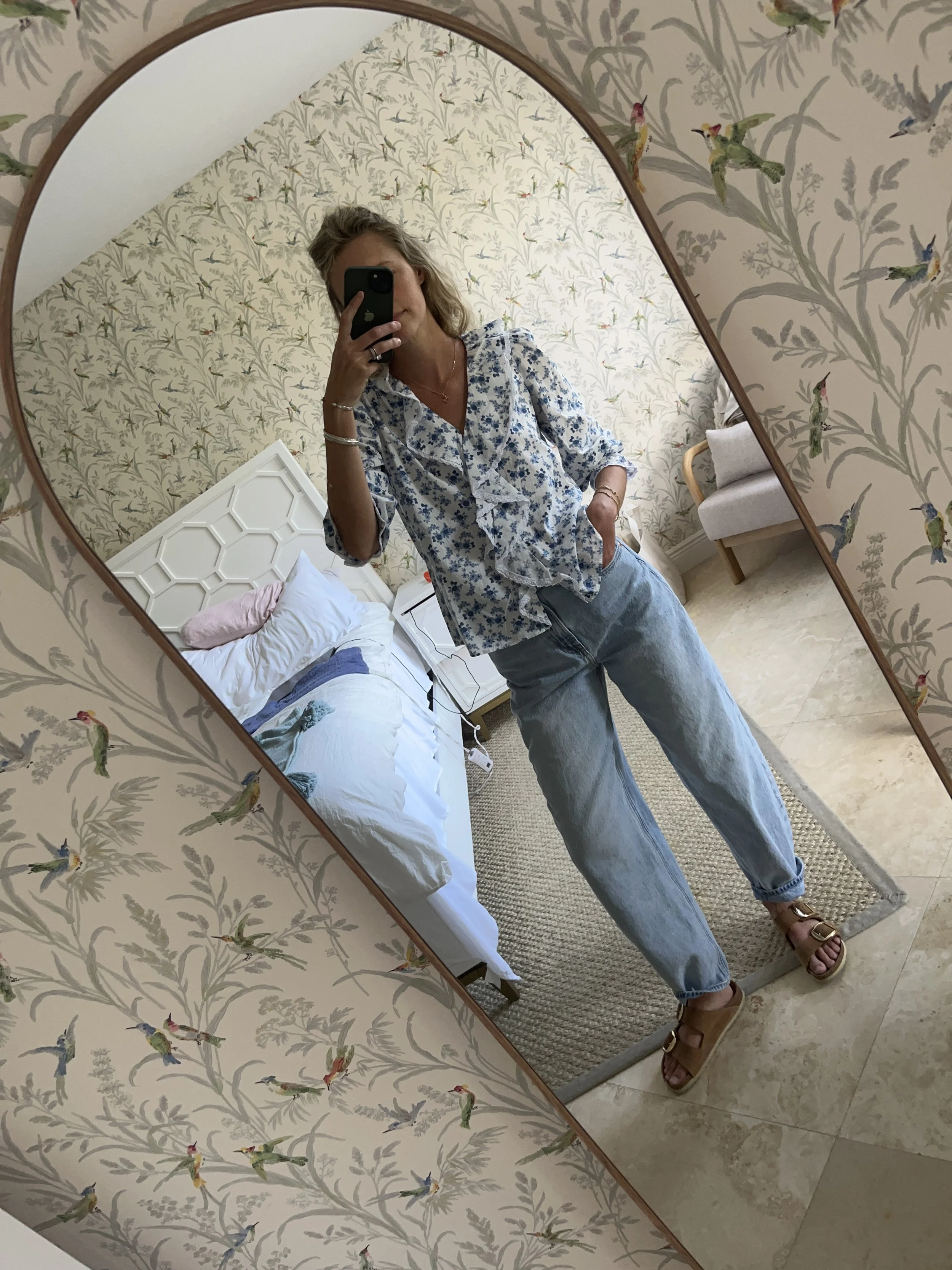 A woman taking a mirror selfie in a spacious living room with wooden floors, large windows, and minimal furniture, wearing a beige jacket, black top, blue jeans, and tan shoes.