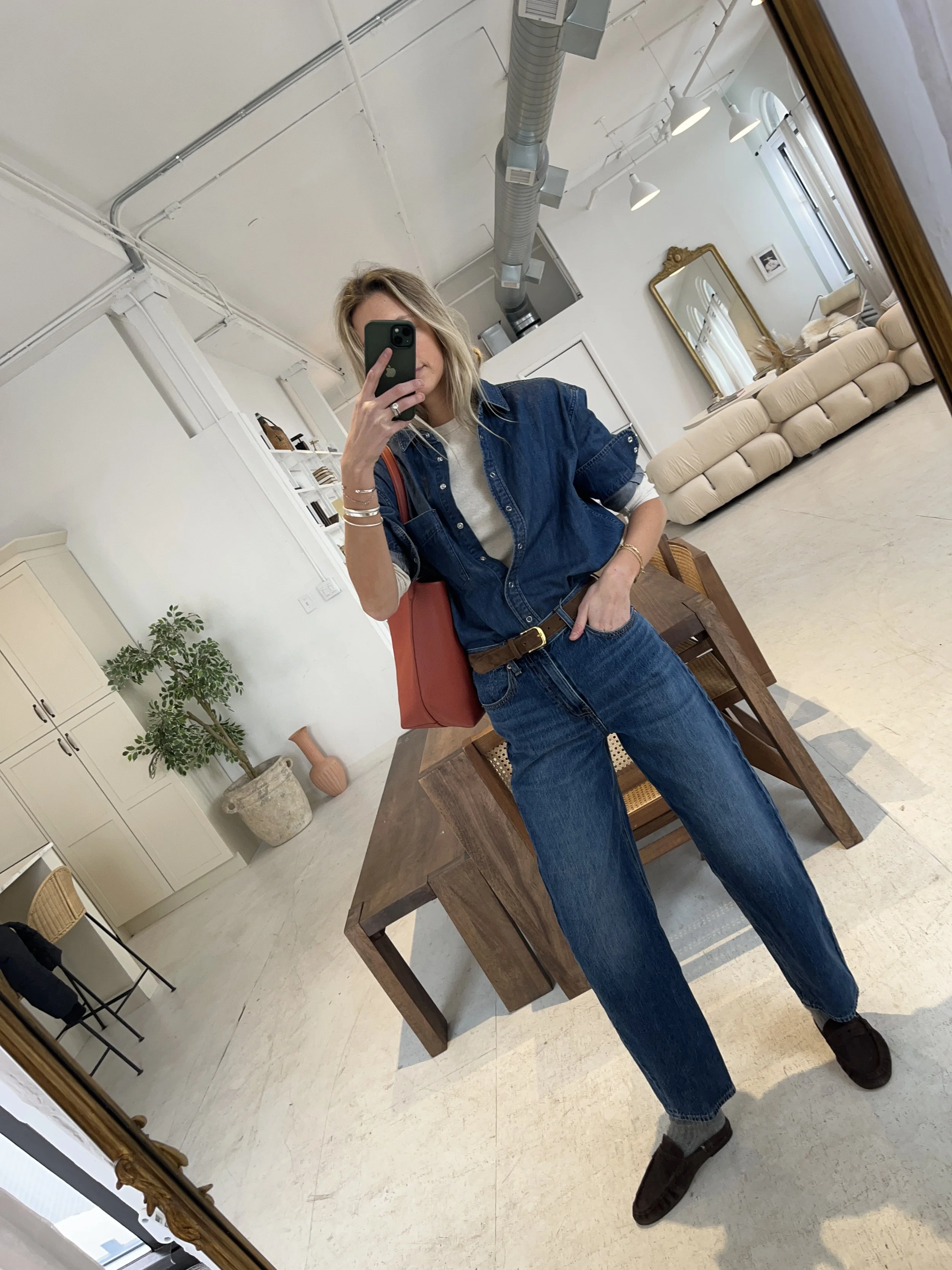 Person taking a mirror selfie in a living room with hardwood floors, wearing a black fleece jacket, gray shirt, black leggings, gray sneakers, and a white cap with a logo.