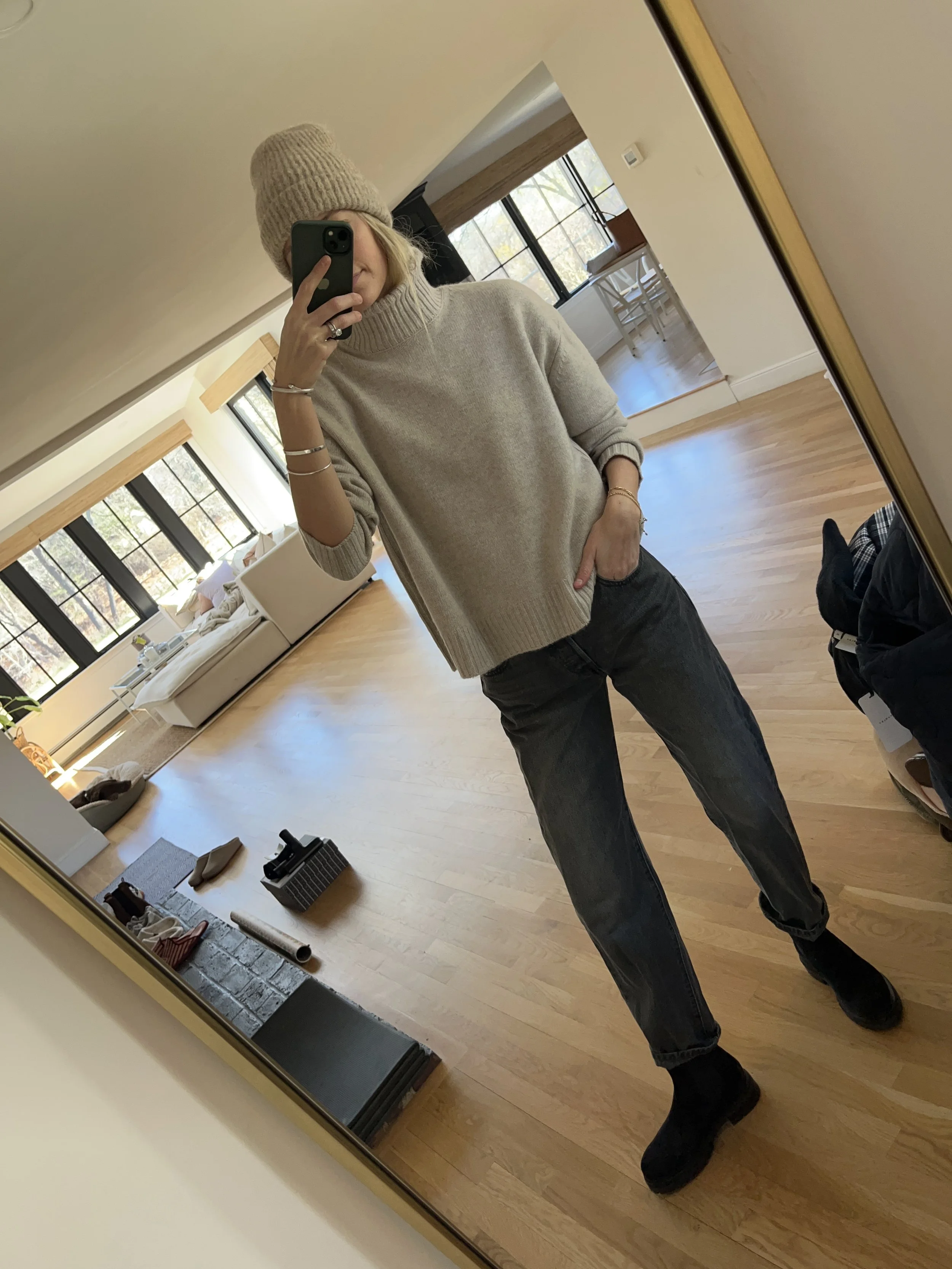 Person taking a mirror selfie in a living room with hardwood floors, wearing a black fleece jacket, gray shirt, black leggings, gray sneakers, and a white cap with a logo.