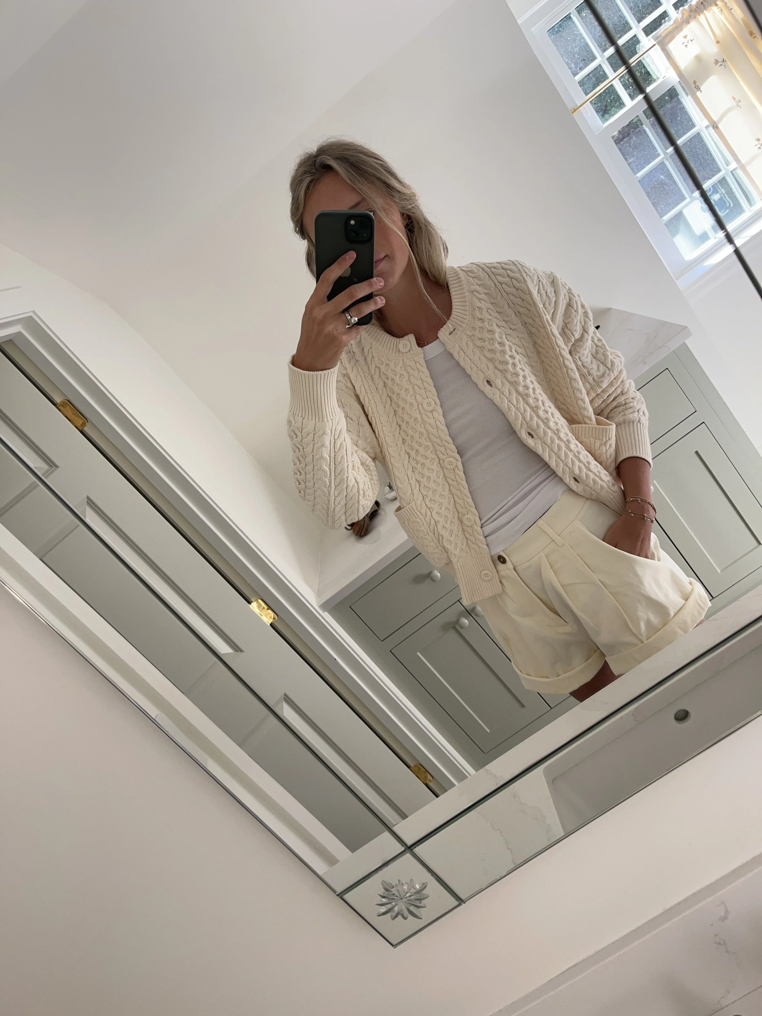 Person taking a mirror selfie in a living room with hardwood floors, wearing a black fleece jacket, gray shirt, black leggings, gray sneakers, and a white cap with a logo.