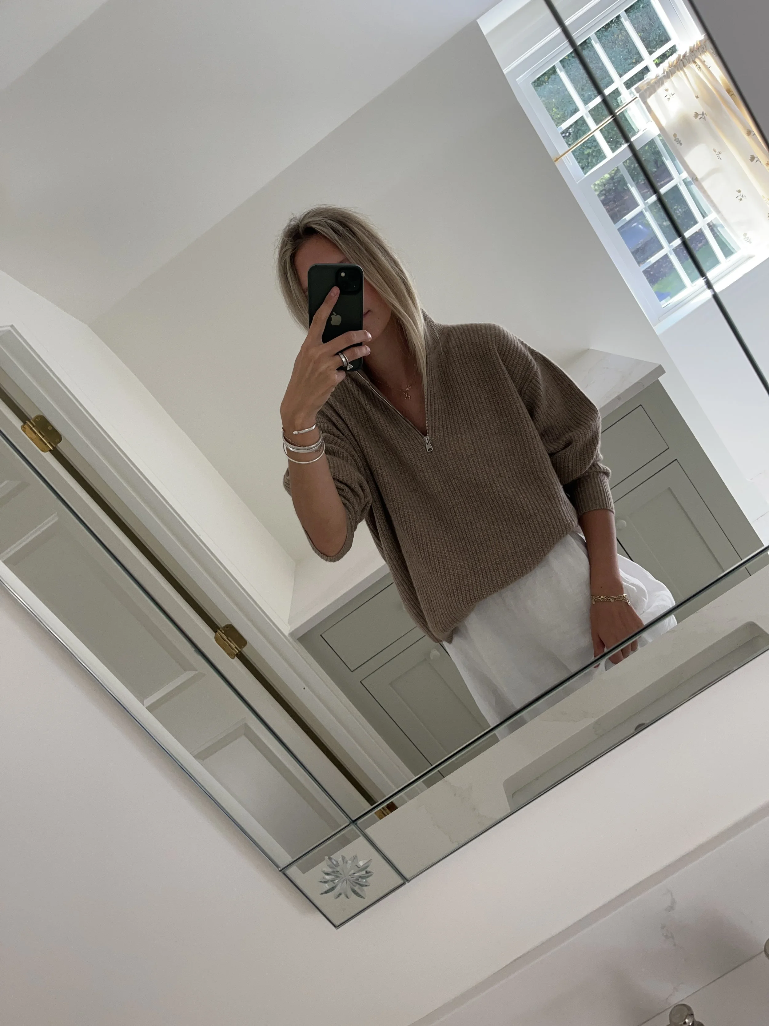Person taking a mirror selfie in a living room with hardwood floors, wearing a black fleece jacket, gray shirt, black leggings, gray sneakers, and a white cap with a logo.